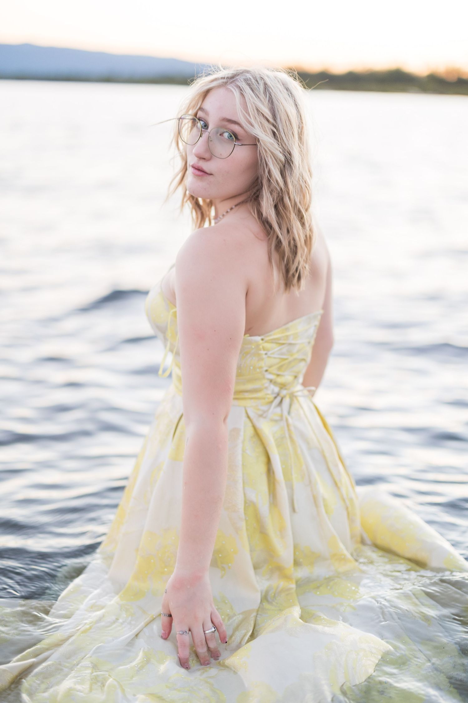 Blonde woman in a yellow floral strapless dress standing in a lake at sunset.