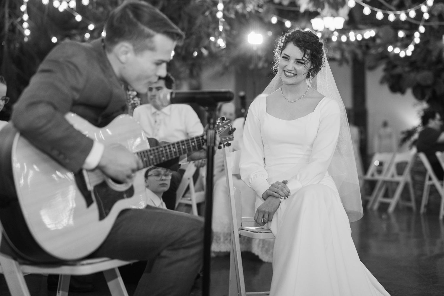 Groom playing guitar for smiling bride at reception.