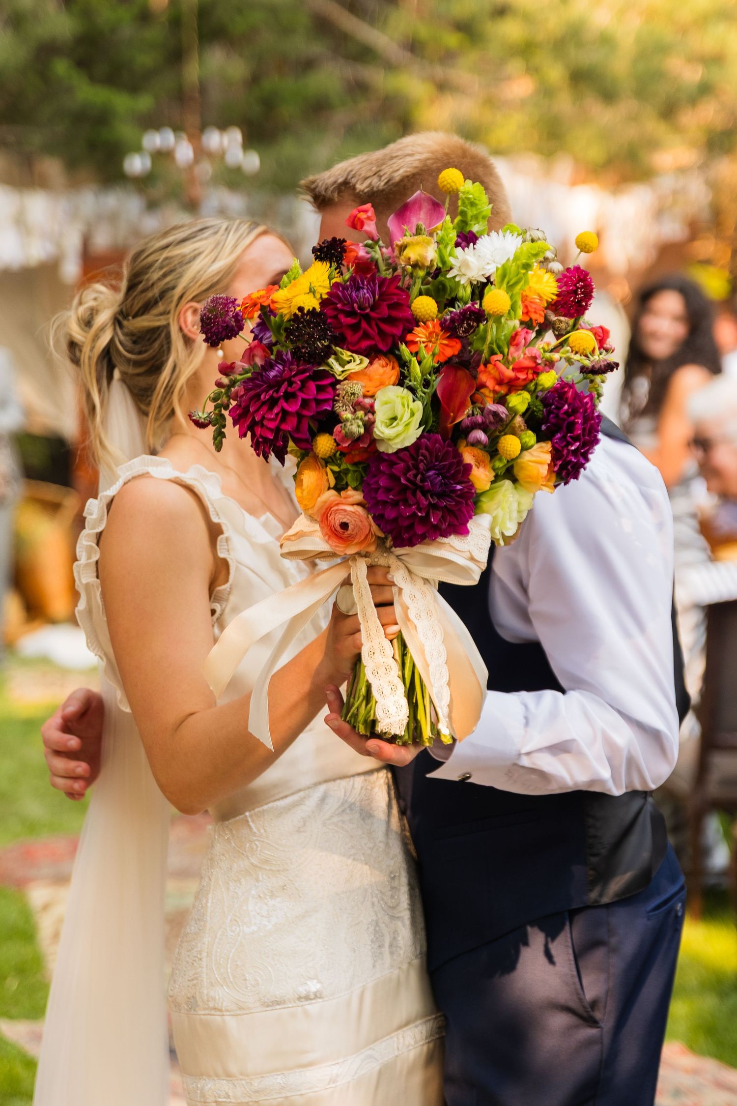 Bride and groom kissing behind a vibrant bouquet of colorful fall flowers at an outdoor wedding reception.