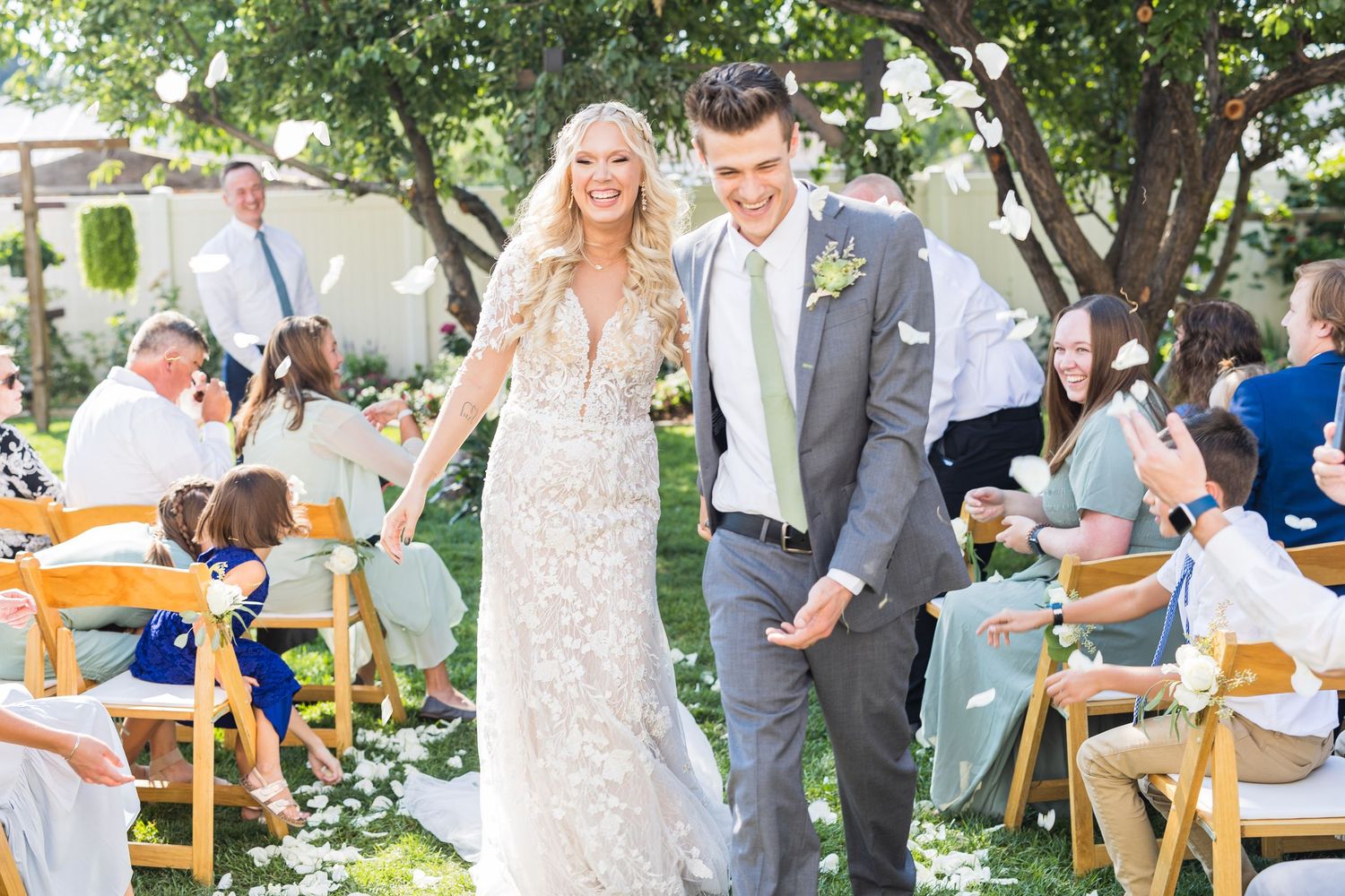 Smiling bride in lace gown and groom in gray suit walk down aisle as guests celebrate at outdoor wedding.
