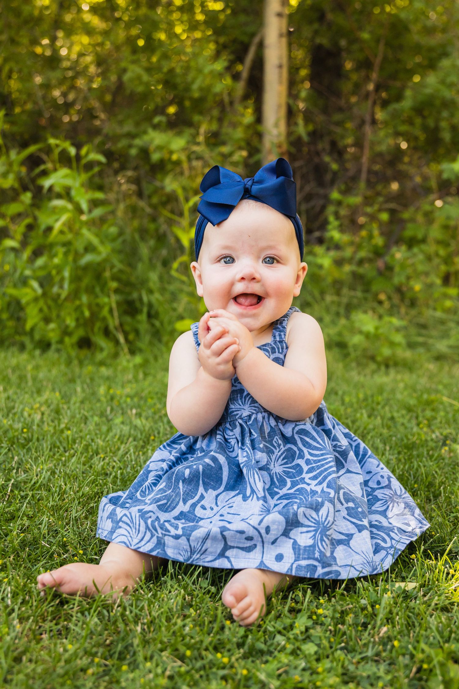 Happy baby girl in blue floral dress with navy bow sitting on green grass outdoors.