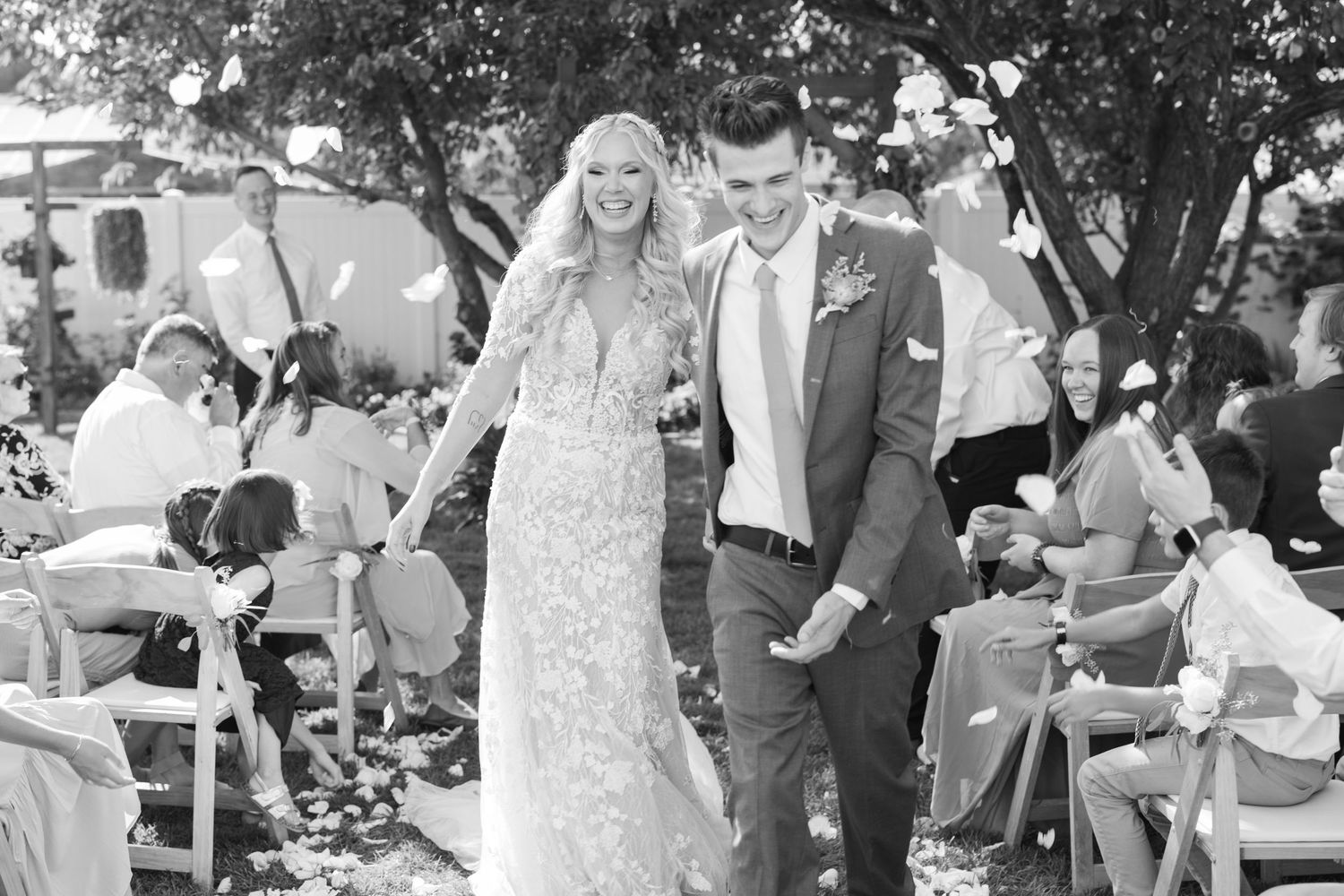Black and white image of joyful bride and groom recessional with guests tossing petals at outdoor wedding.