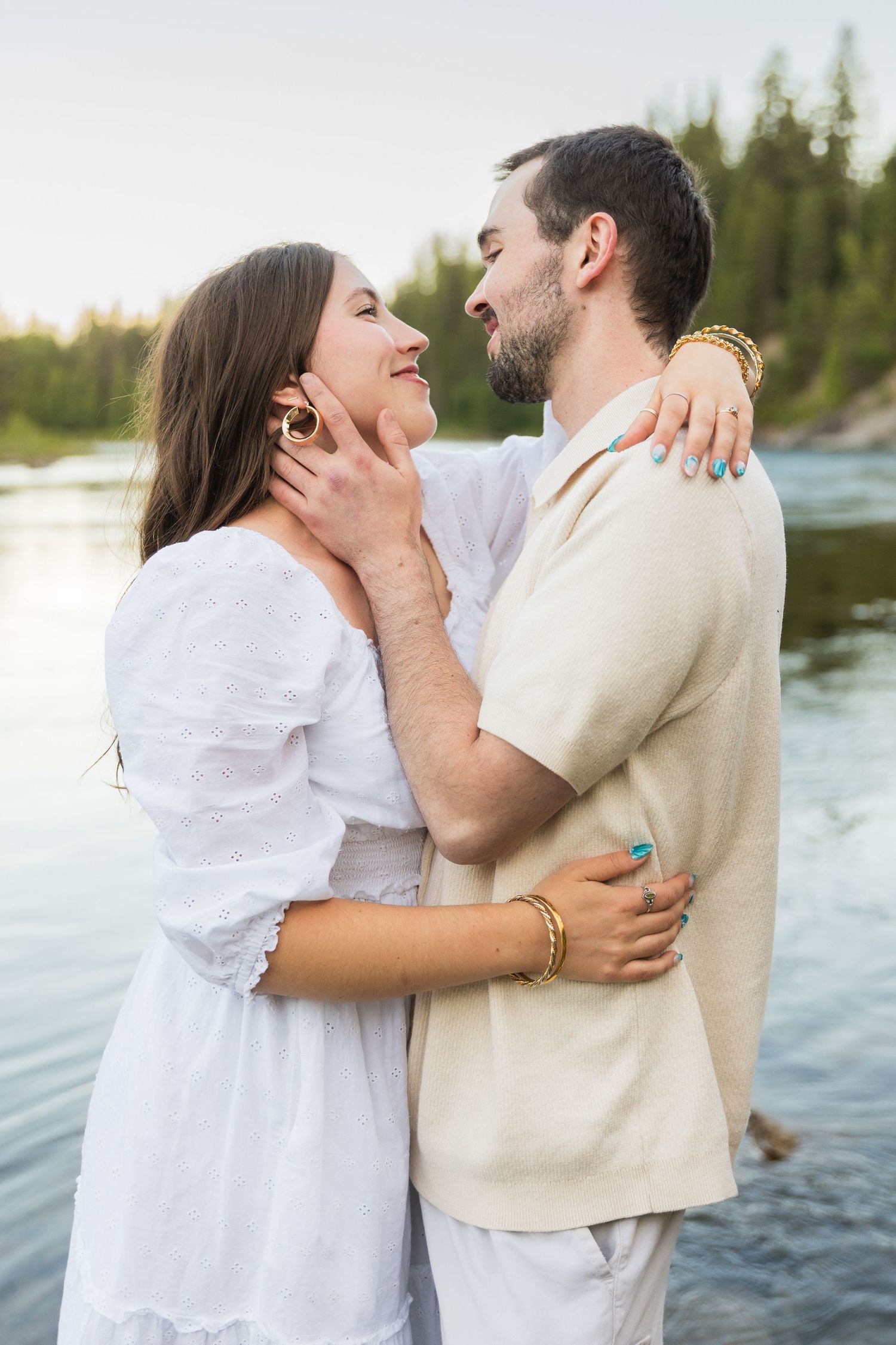 Couple embracing and smiling at each other by a river, woman in white dress, man in cream shirt, surrounded by trees.