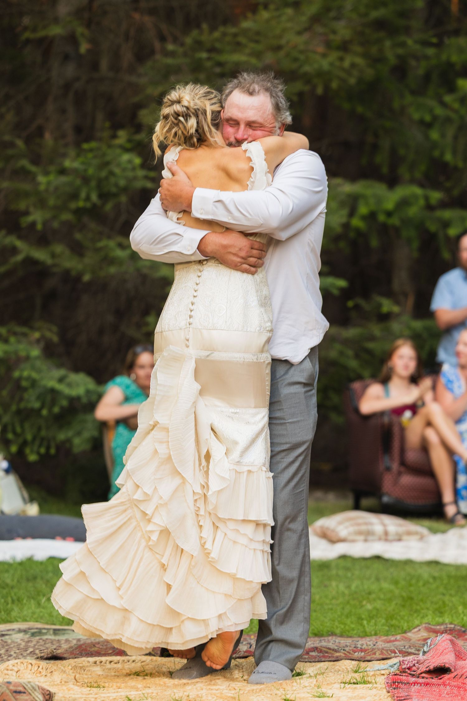 Bride in ruffled ivory wedding dress shares emotional dance with older man at outdoor woodland wedding reception.