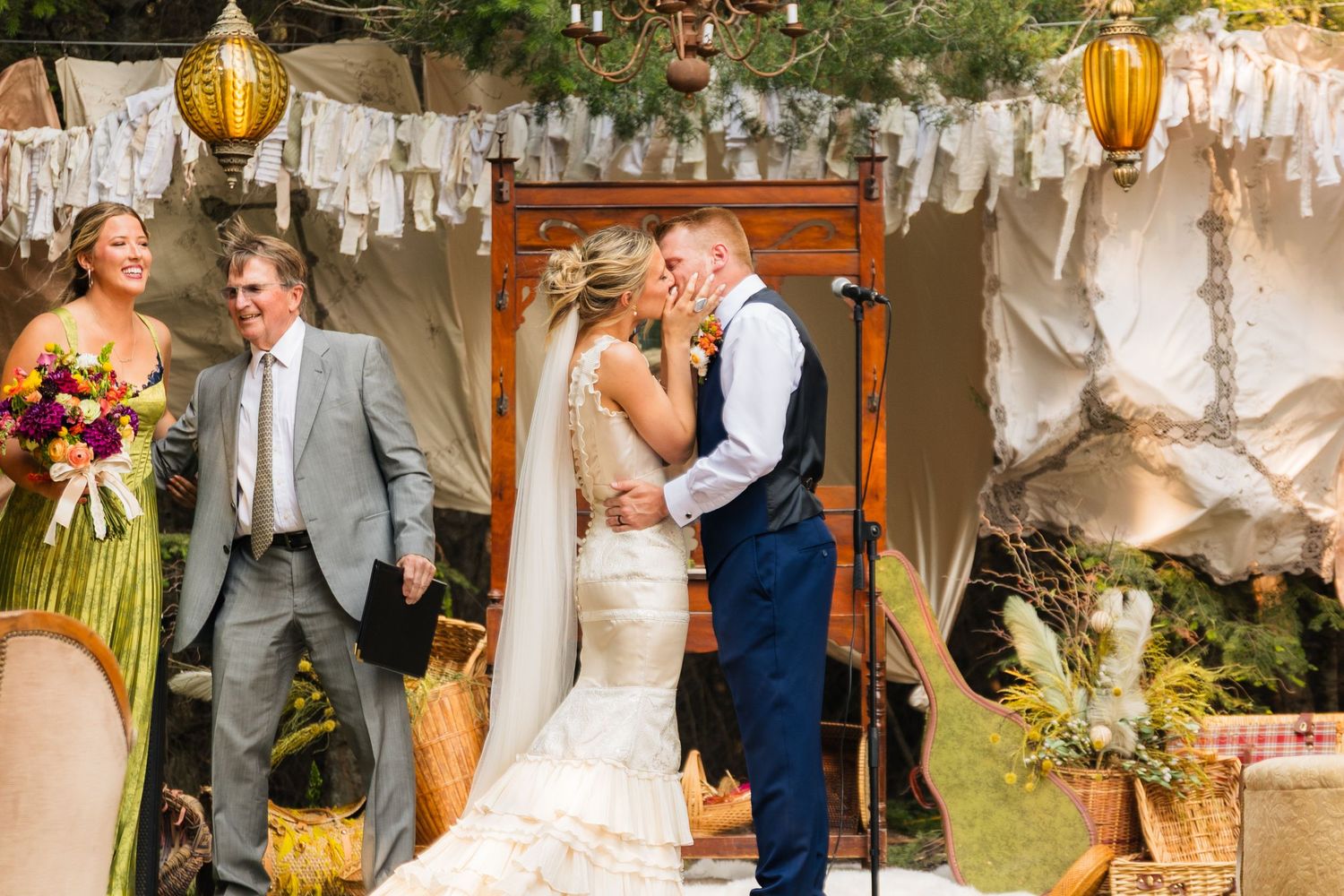 Bride and groom share first kiss at outdoor boho wedding ceremony with floral decor and tent backdrop.