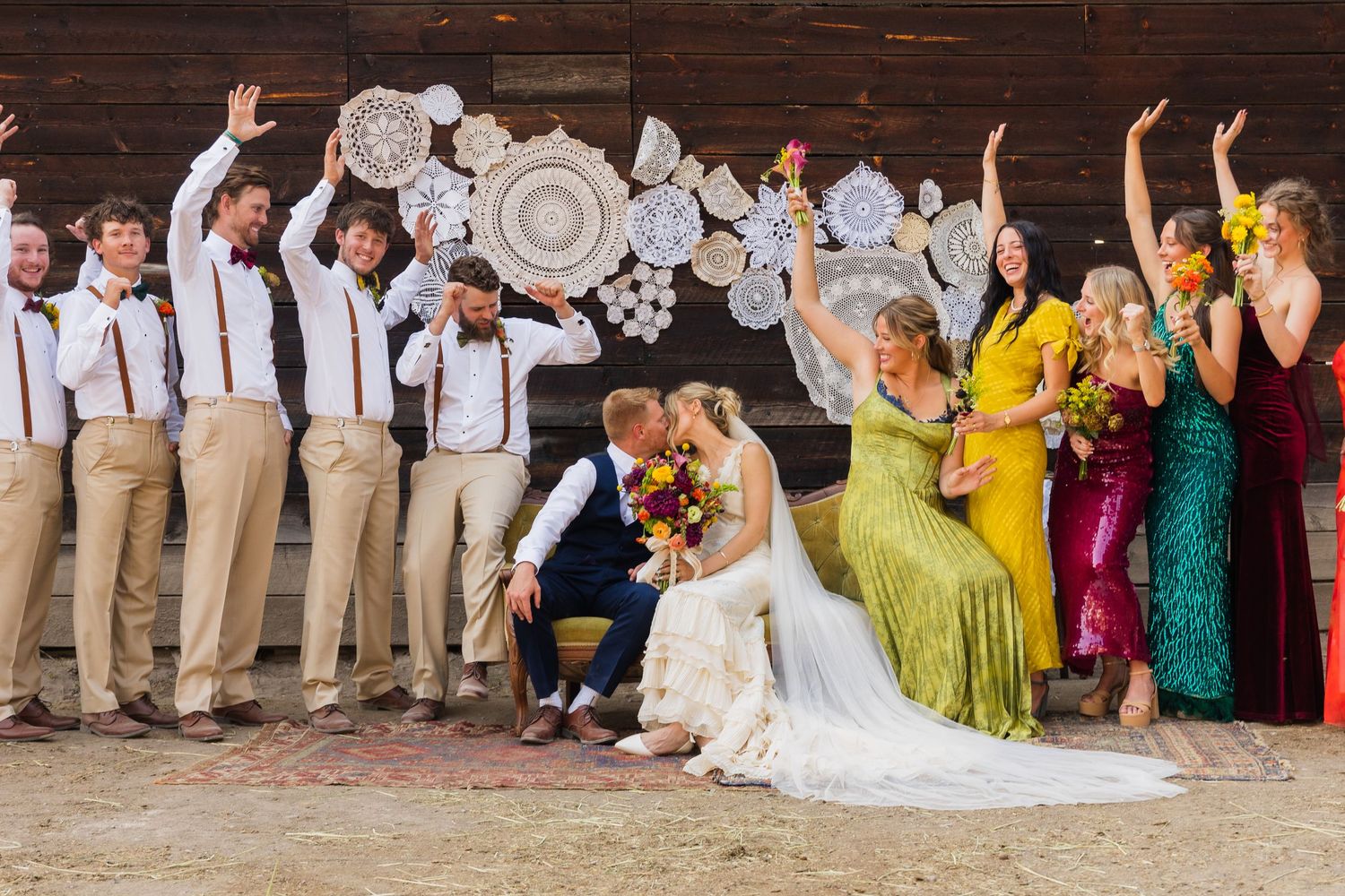 Wedding party celebrating with raised arms beside kissing bride and groom in front of a barn.