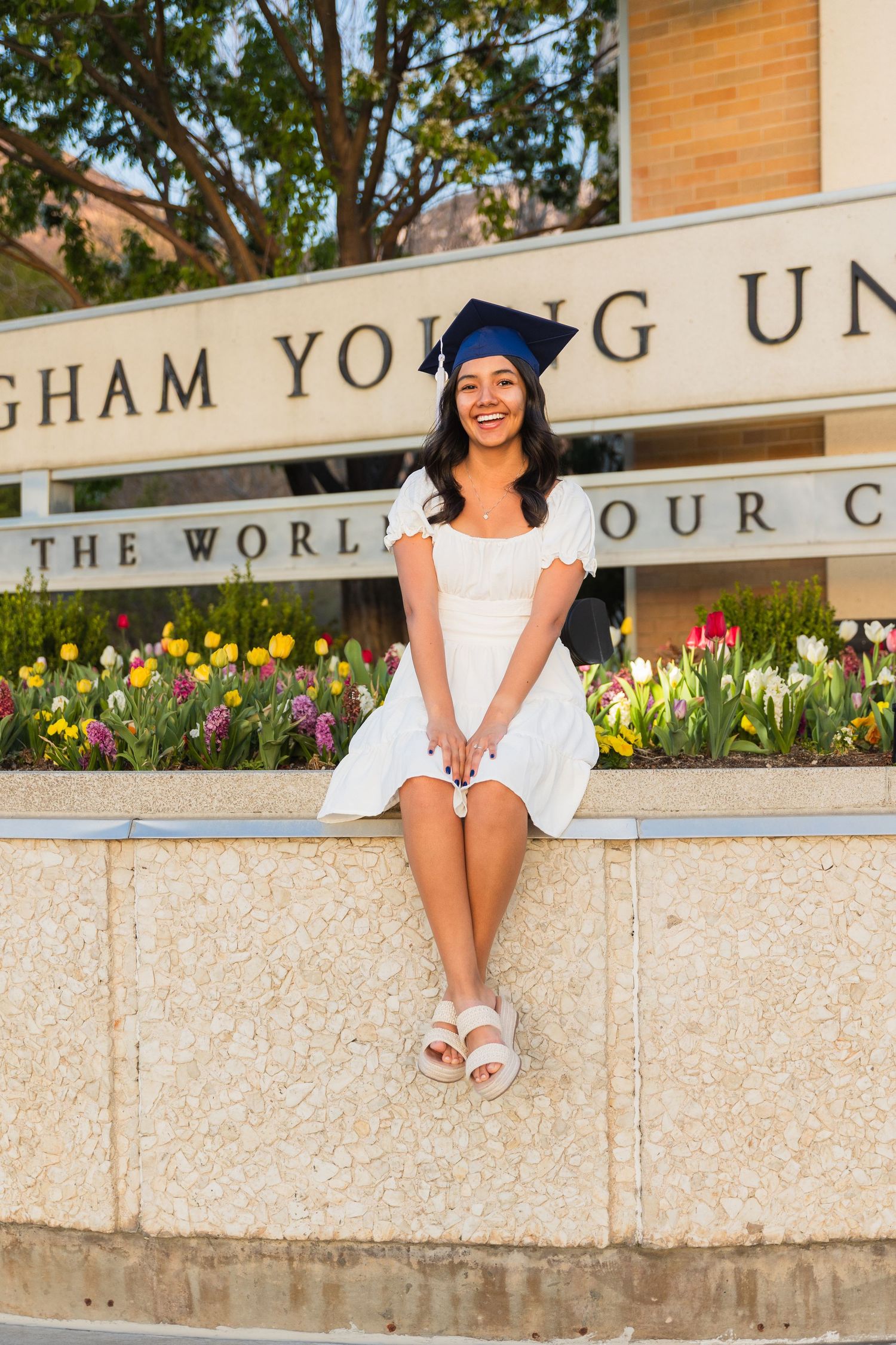 Graduate in cap and white dress sitting at Brigham Young University sign with tulips.