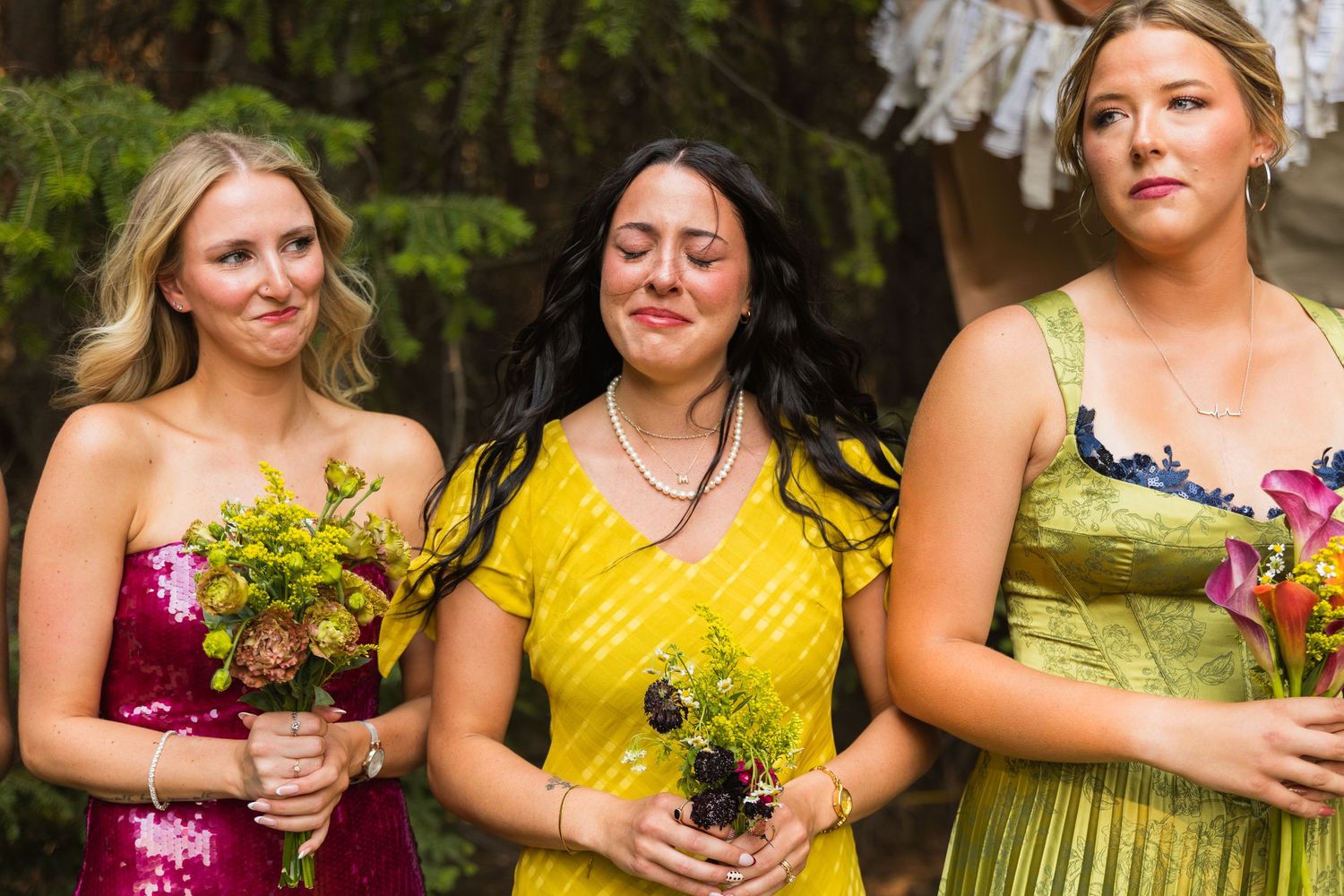 Three bridesmaids in colorful dresses holding bouquets with emotional expressions at a wedding ceremony.