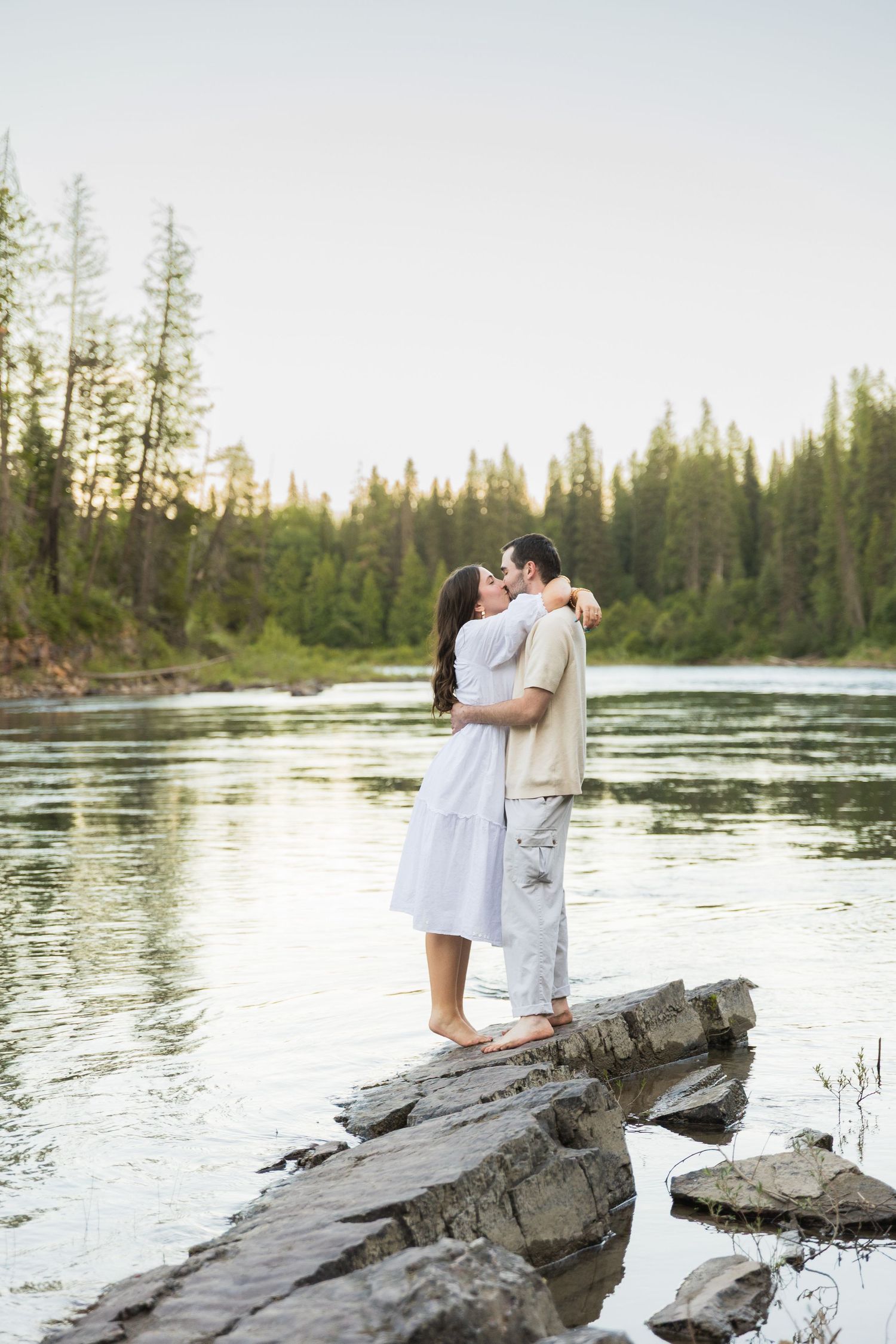 Couple embracing and kissing on rocky shoreline beside calm river surrounded by tall evergreen trees at sunset.