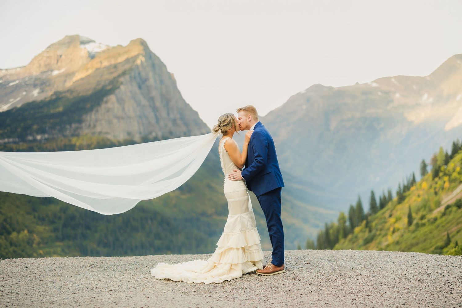 Bride and groom kissing with veil blowing in wind, stunning mountain backdrop at Glacier National Park wedding.