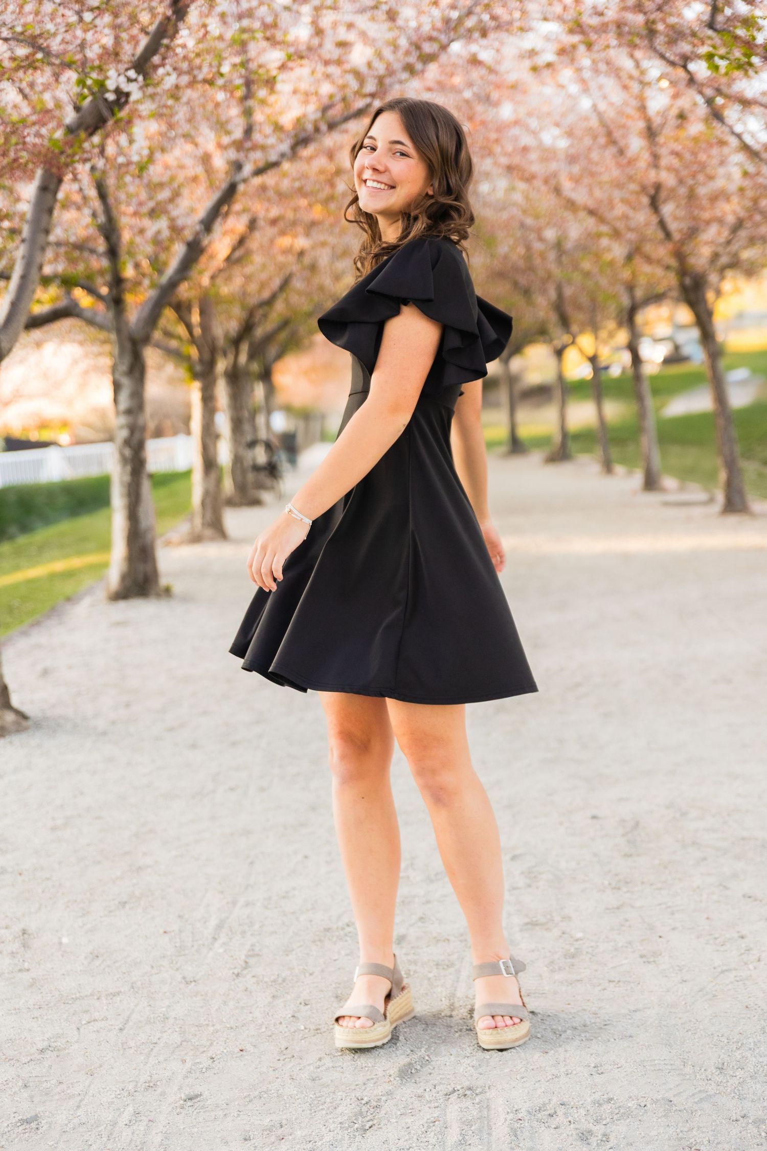 Young woman in a black ruffled dress smiling in a cherry blossom tree-lined path.