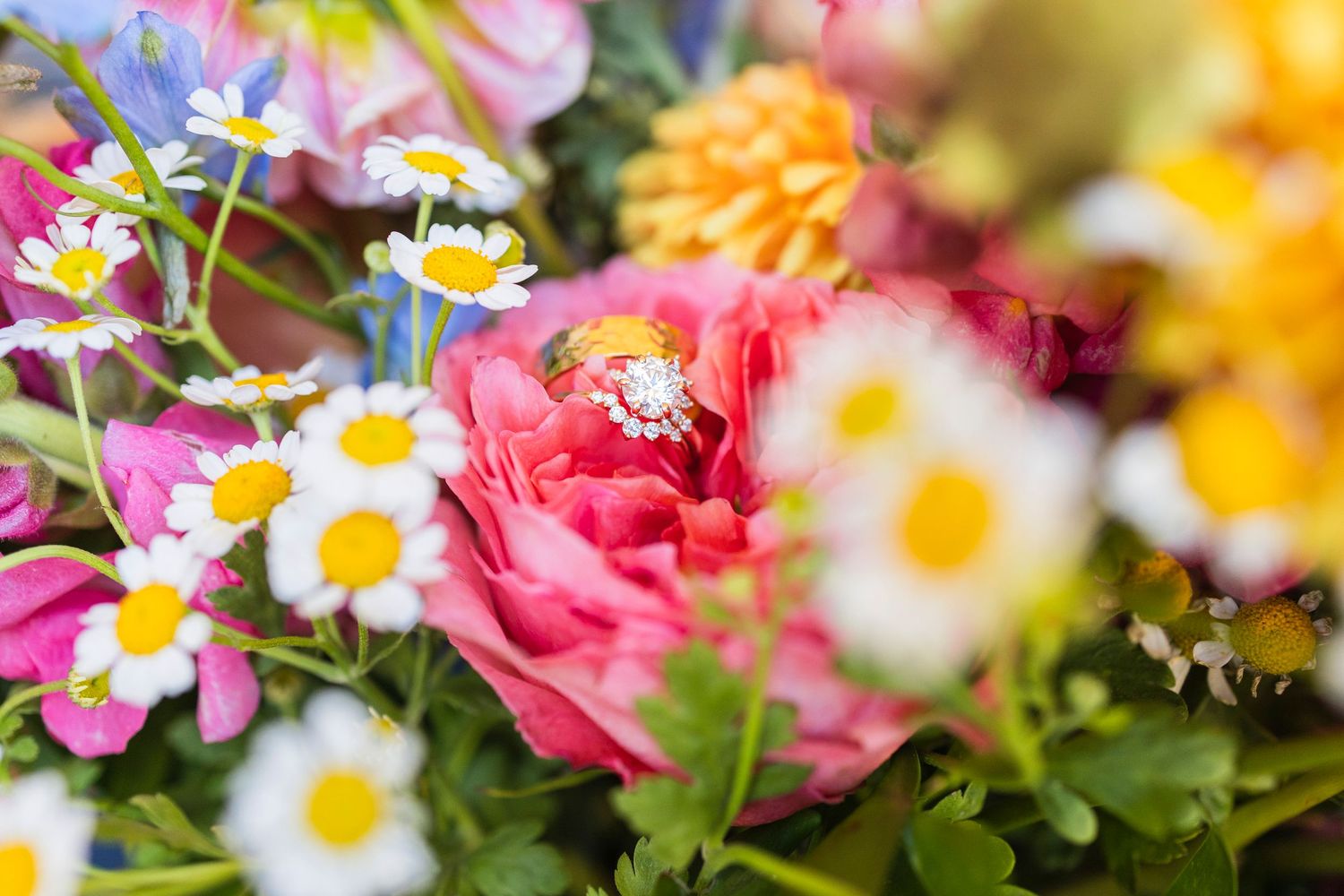 Close-up of a diamond engagement ring nestled among colorful wedding bouquet flowers including roses and daisies.