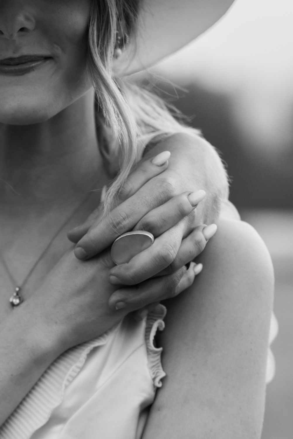 Black and white photo of a woman in a hat wearing a ring, with hands crossed over her shoulder.