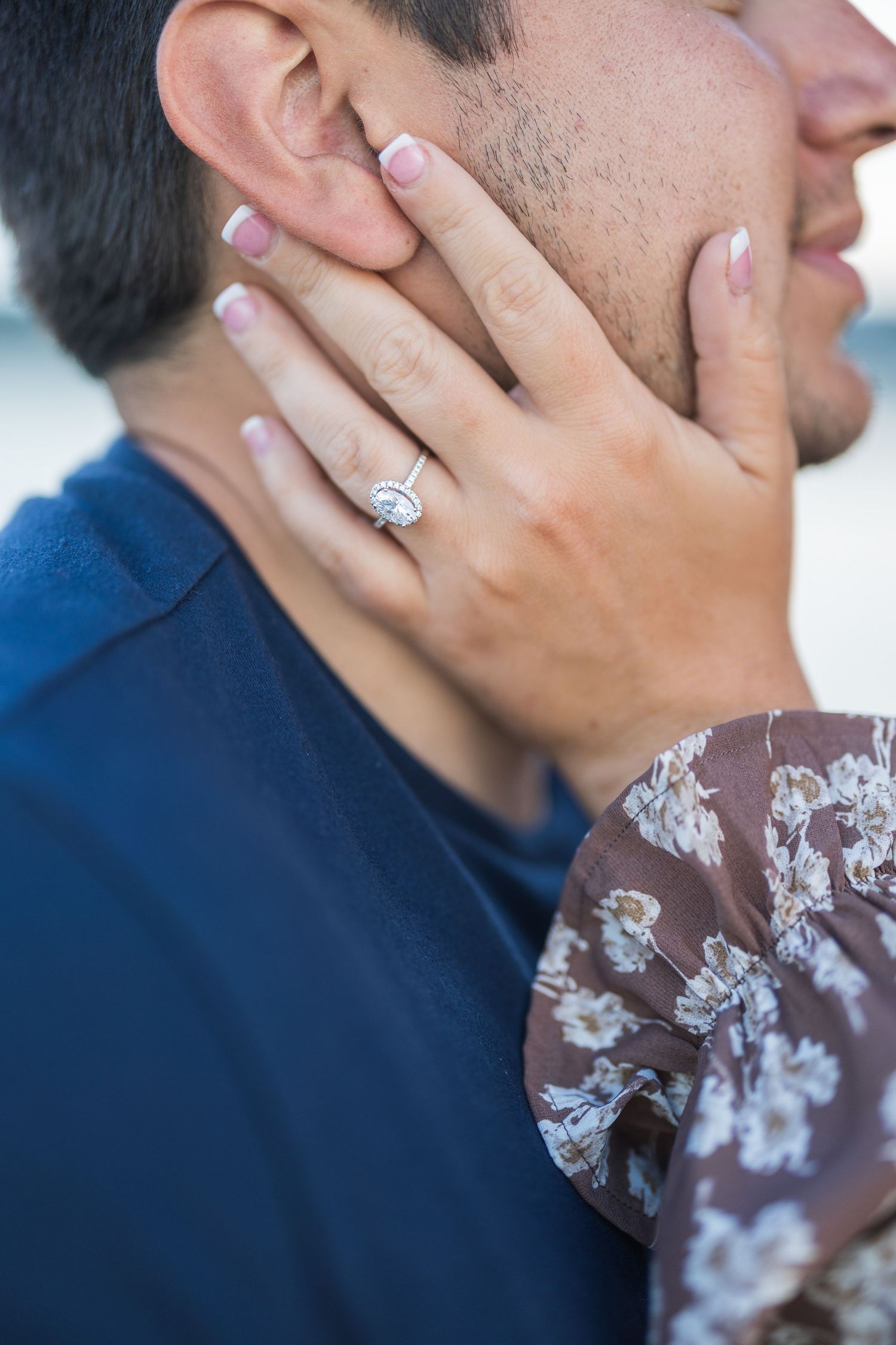 Close-up of womans hand with cushion-cut halo diamond engagement ring resting on mans face.