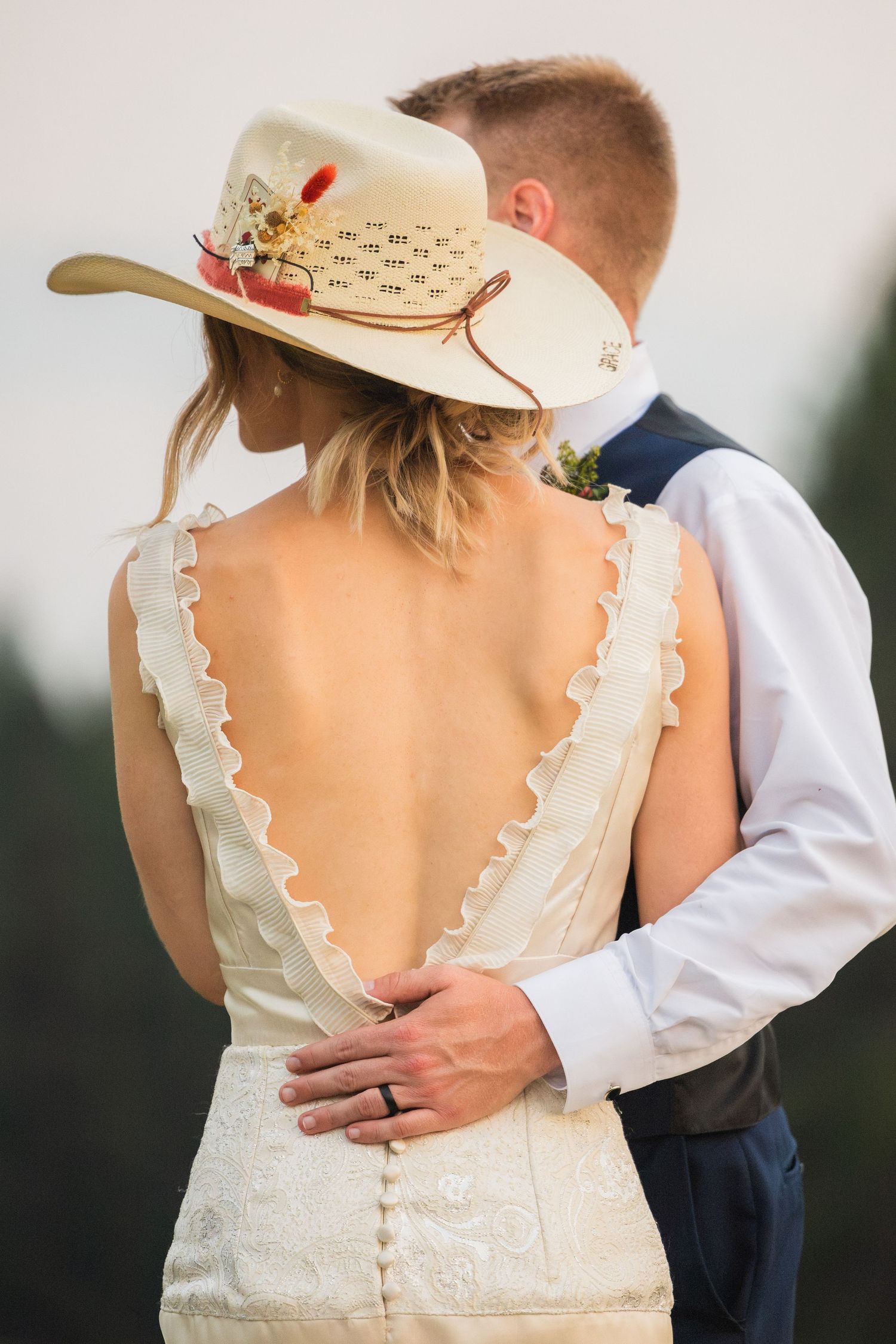Bride in backless lace wedding dress and cowboy hat embraced by groom in vest at outdoor wedding.