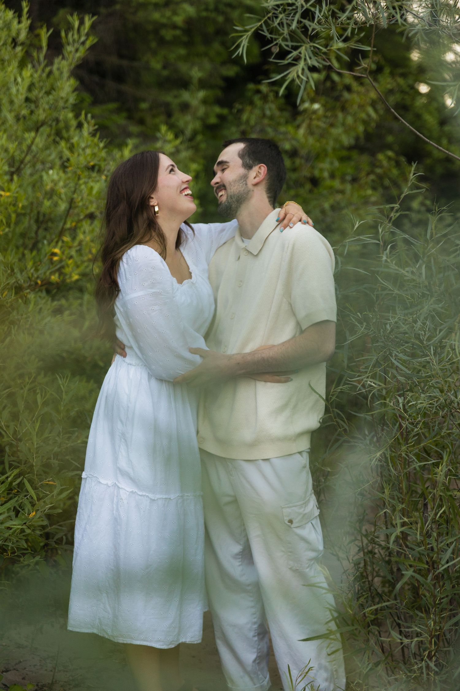 A couple in white outfits embracing and smiling at each other surrounded by lush green foliage outdoors.