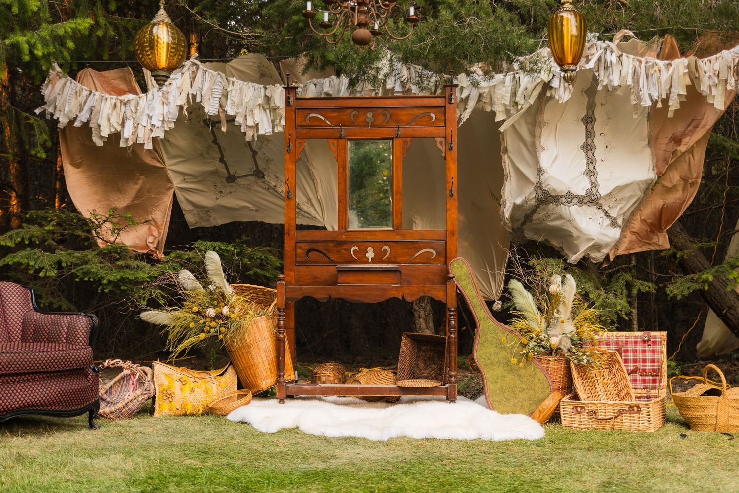 Rustic outdoor boho wedding setup with vintage wooden armoire, wicker baskets, draped fabric, and golden lanterns on green lawn.