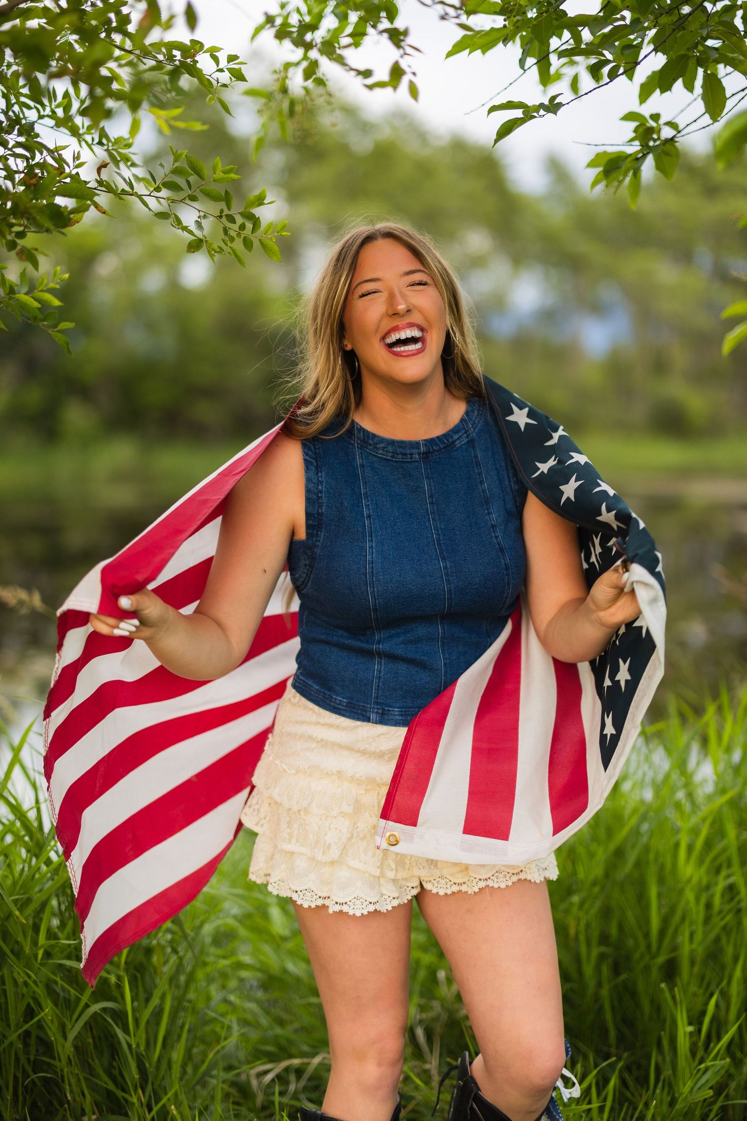 Laughing blonde woman holding American flag outdoors in green nature setting, wearing denim top and white lace shorts.