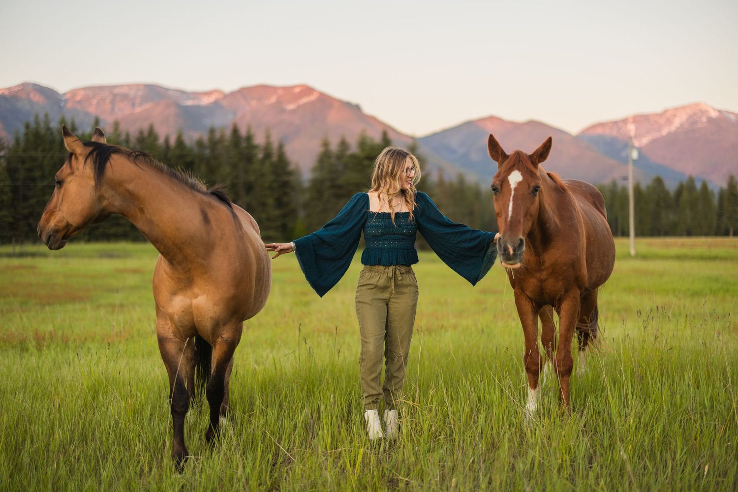 A blonde woman in a teal top stands between two brown horses in a green meadow with mountain backdrop.
