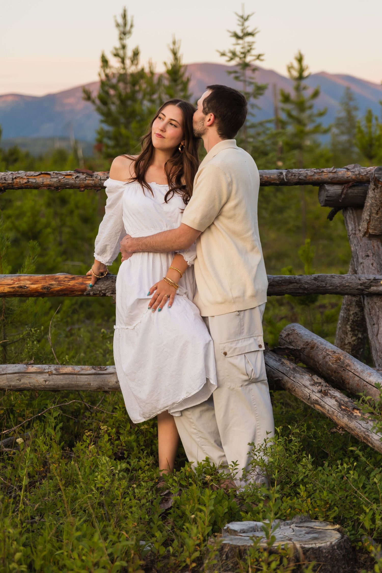 Romantic couple embracing by rustic log fence with mountain and pine tree backdrop at sunset.