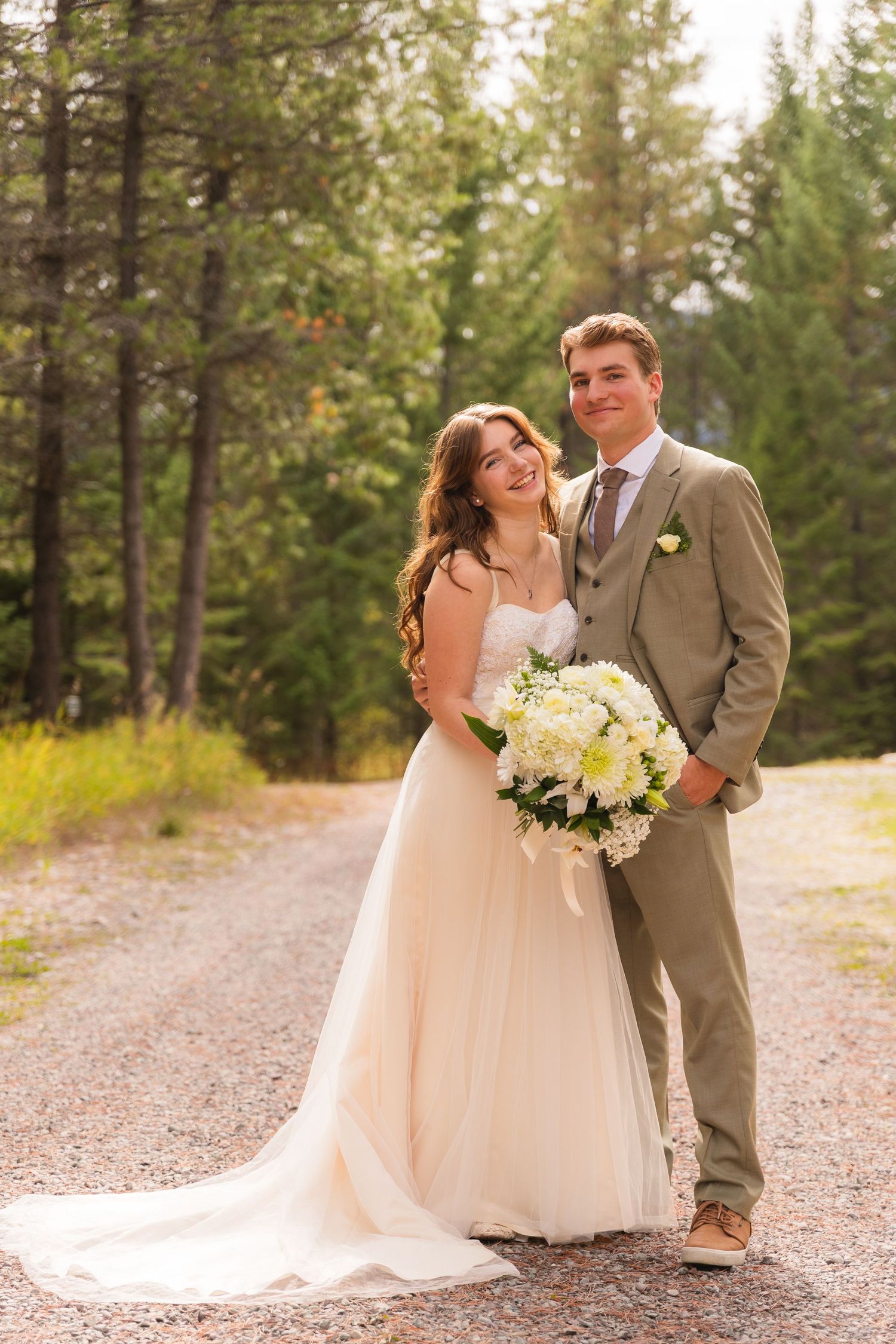 A smiling bride and groom pose on a forest path, she holds white flowers in a flowing gown.