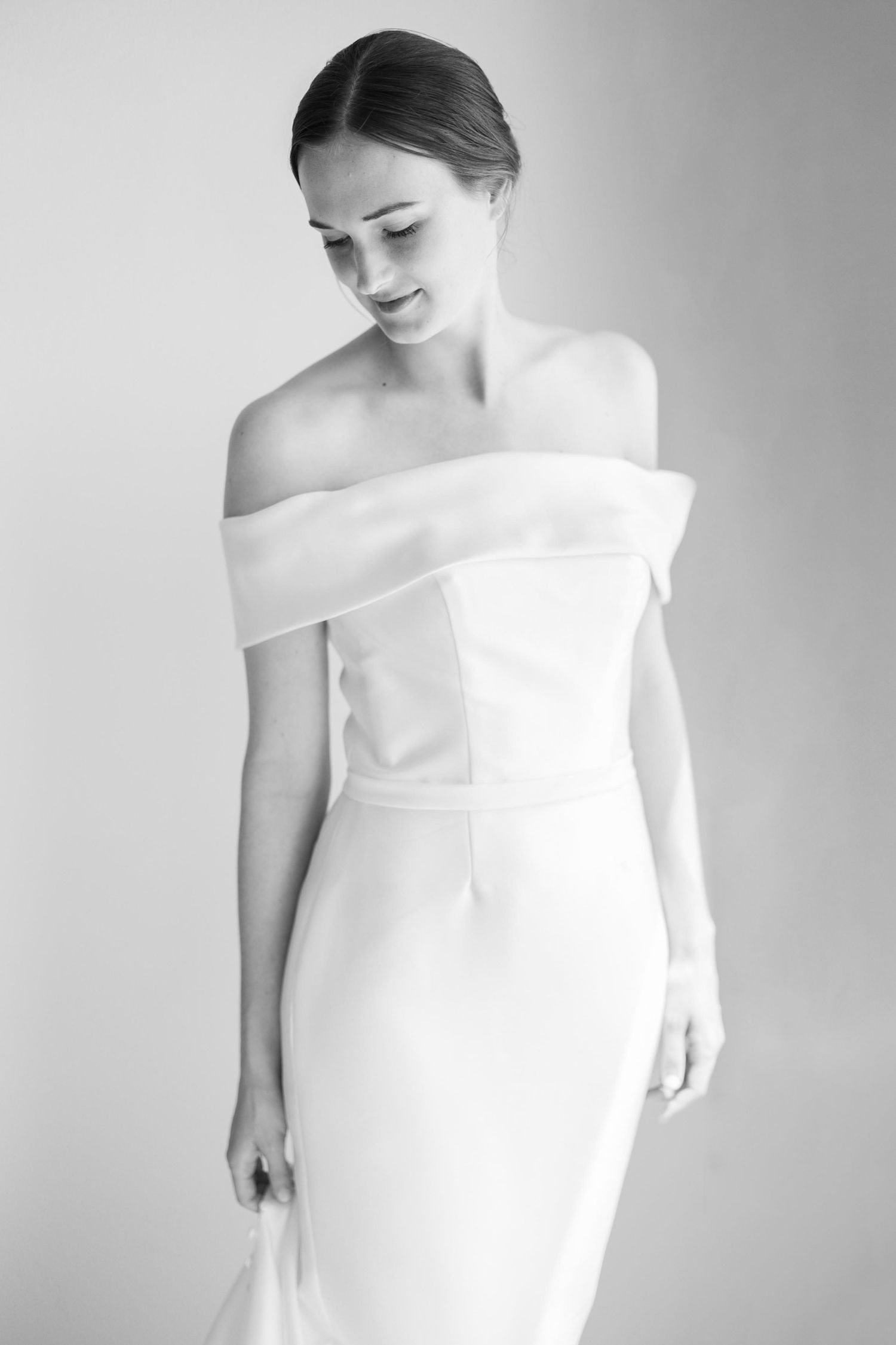 Bride in minimalist off-shoulder white wedding dress poses gracefully in black and white portrait.