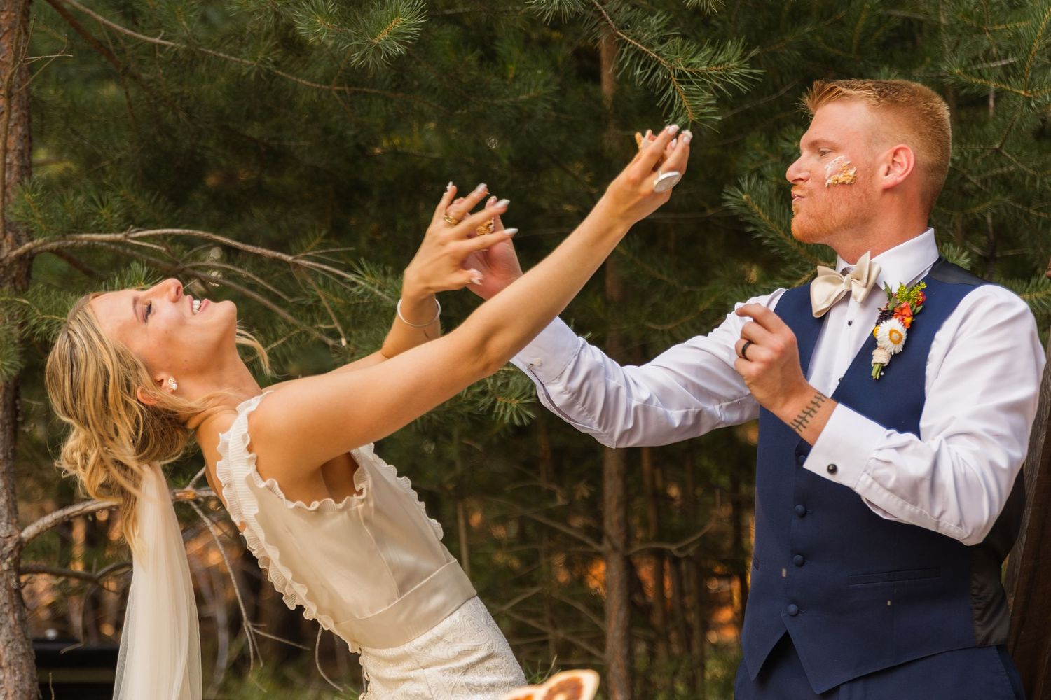 Bride and groom laughing and dancing together outdoors among pine trees during wedding reception.