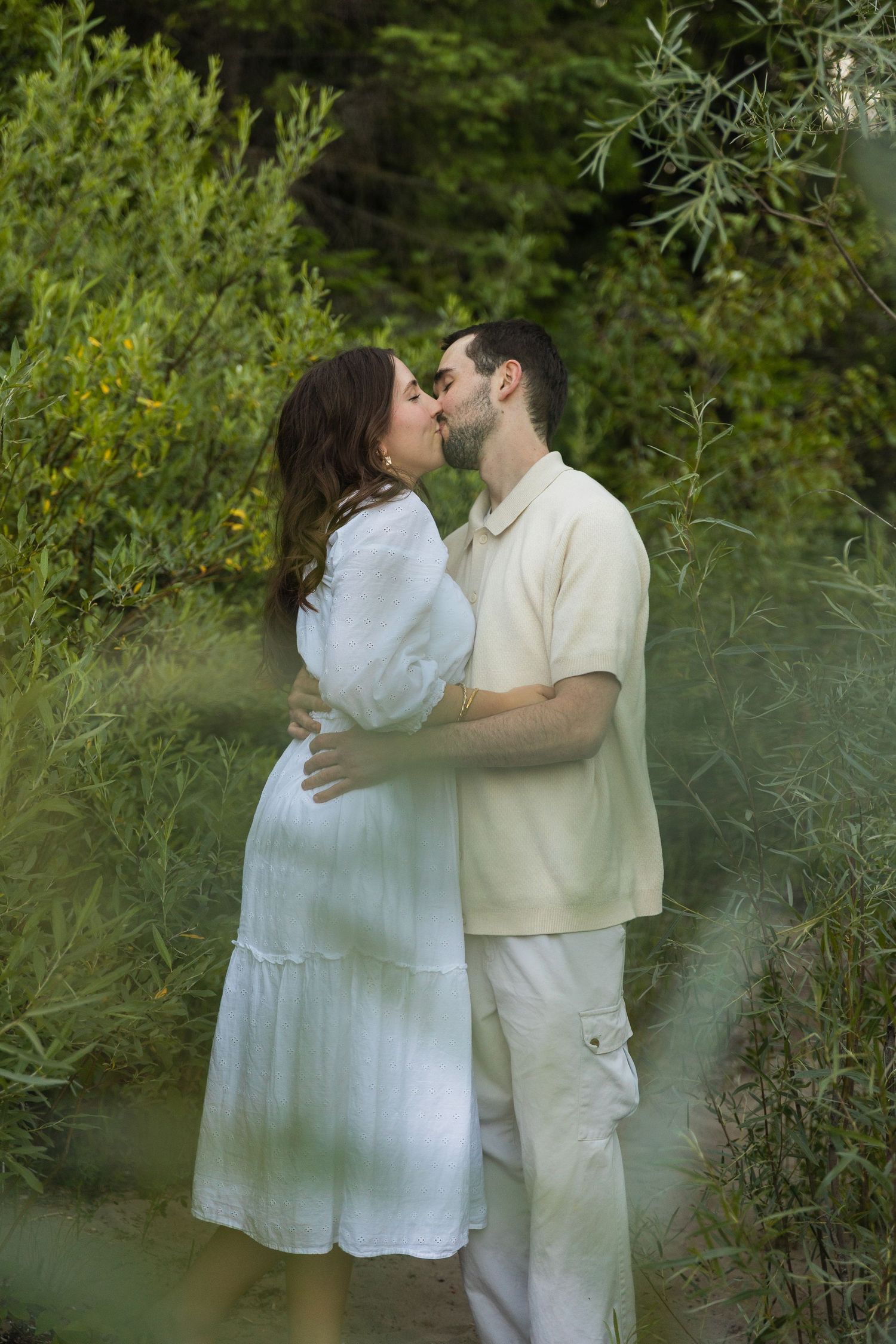 Pregnant woman in white dress kissing partner surrounded by lush green foliage in a garden setting.