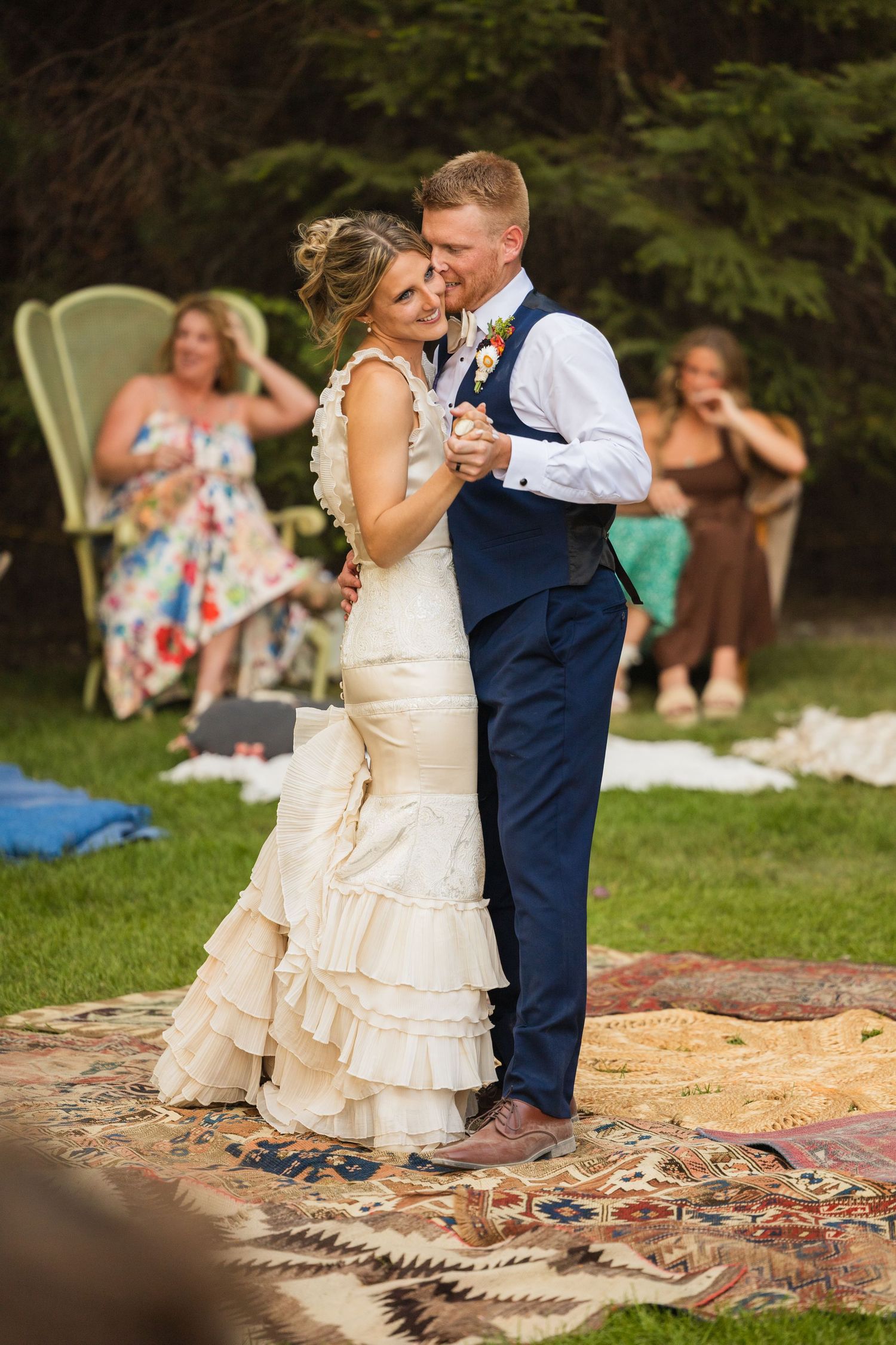 Bride and groom share first dance on a patterned rug outdoors at a rustic backyard wedding with guests watching.