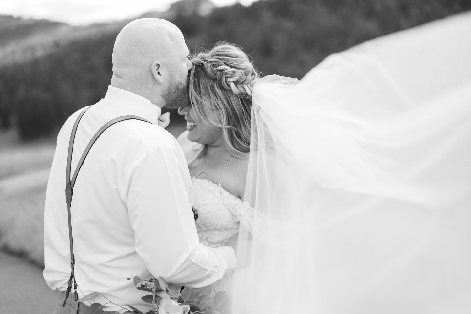 Black and white romantic photo of bald groom kissing bride as her long veil flows in the breeze.