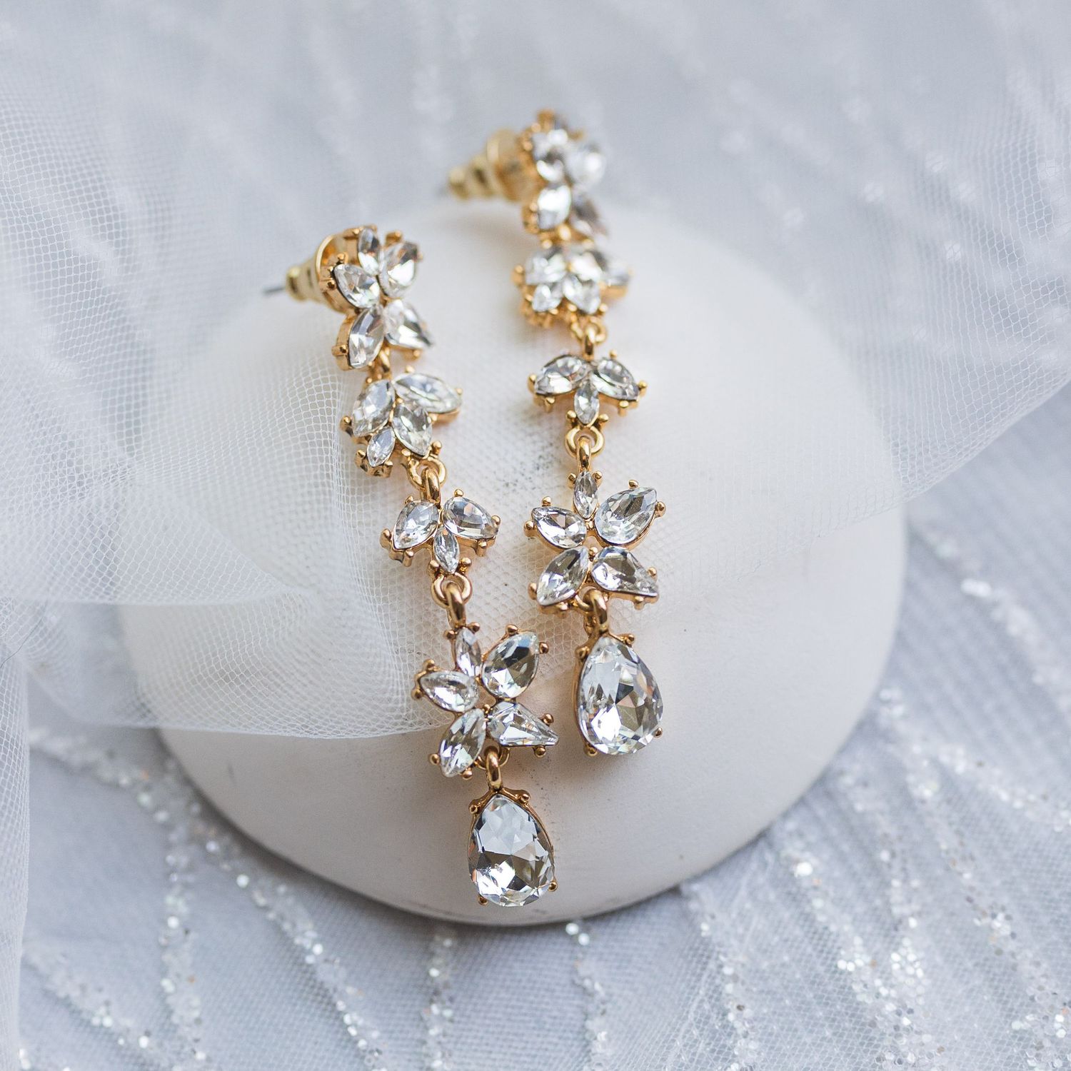 Long gold crystal drop earrings with teardrop pendants displayed on white surface with sheer fabric.