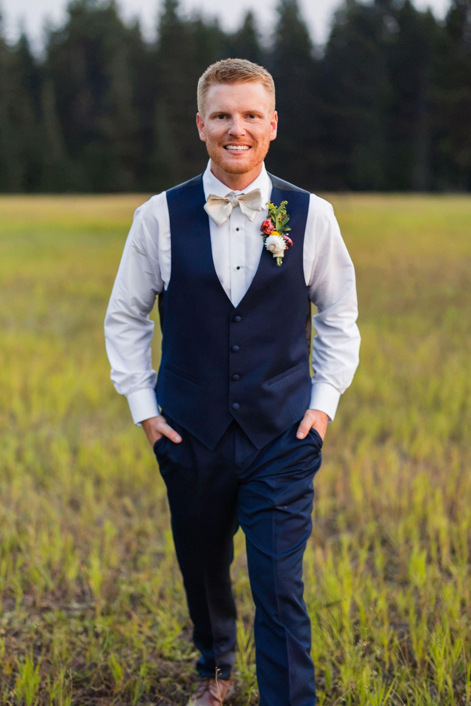 Smiling groom in navy vest, white shirt, and bow tie with boutonniere standing in a green field.