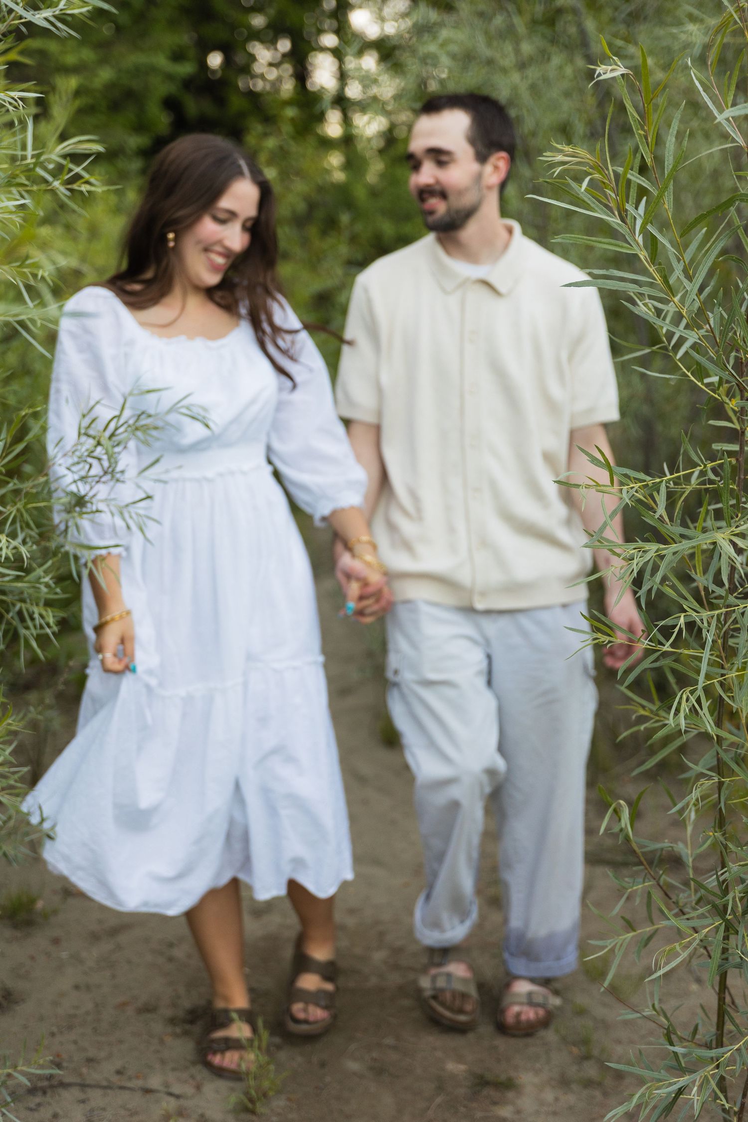 Happy couple holding hands and walking through a tree-lined path, dressed in coordinating white and cream outfits.