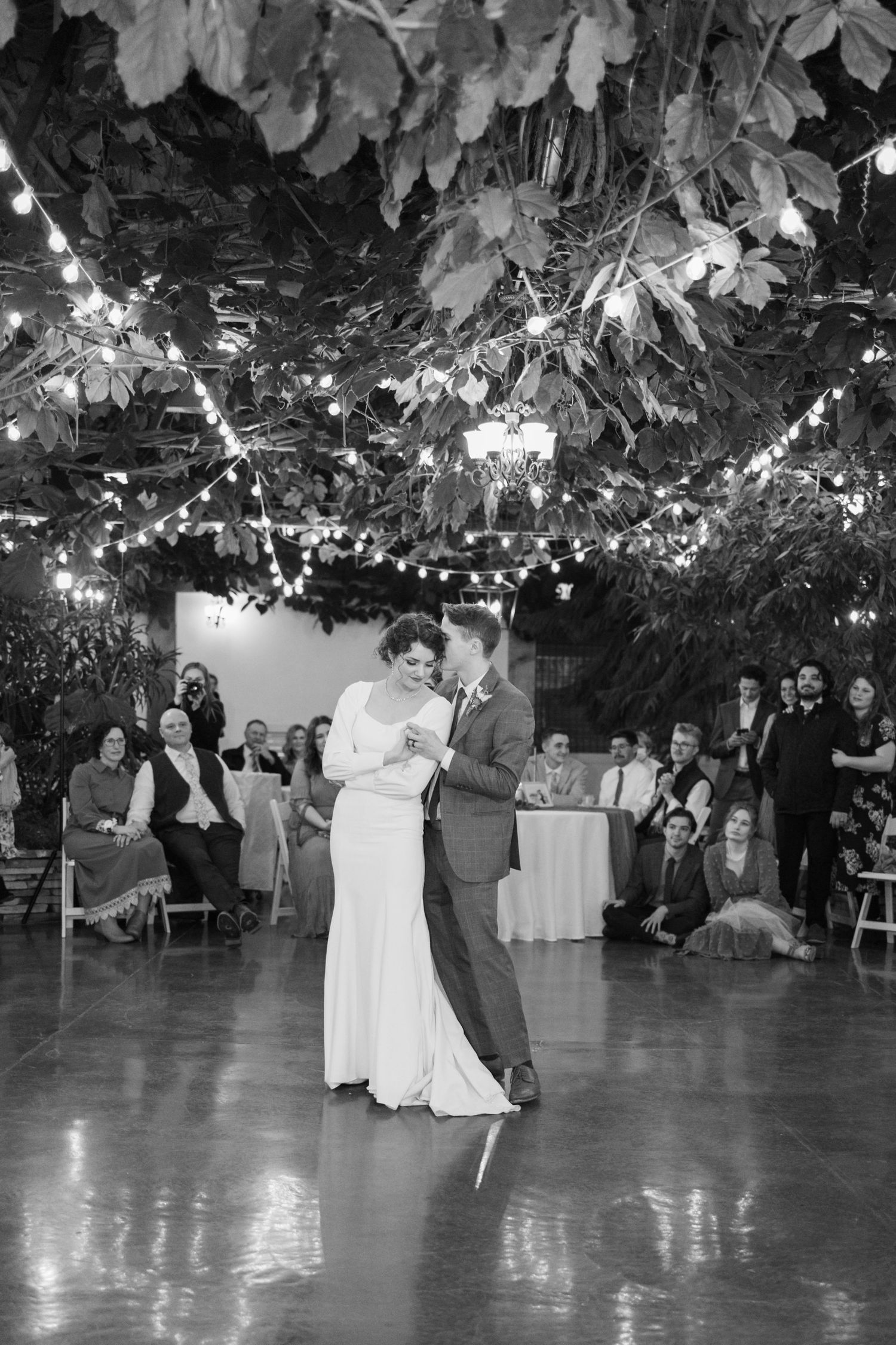 Black and white photo of a couple sharing their first dance under string lights and lush greenery.