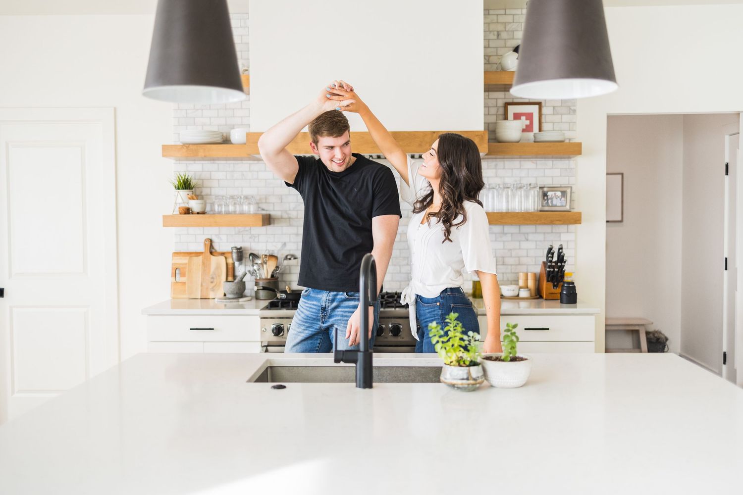 Happy couple dancing together in modern white kitchen with open shelving and pendant lights.