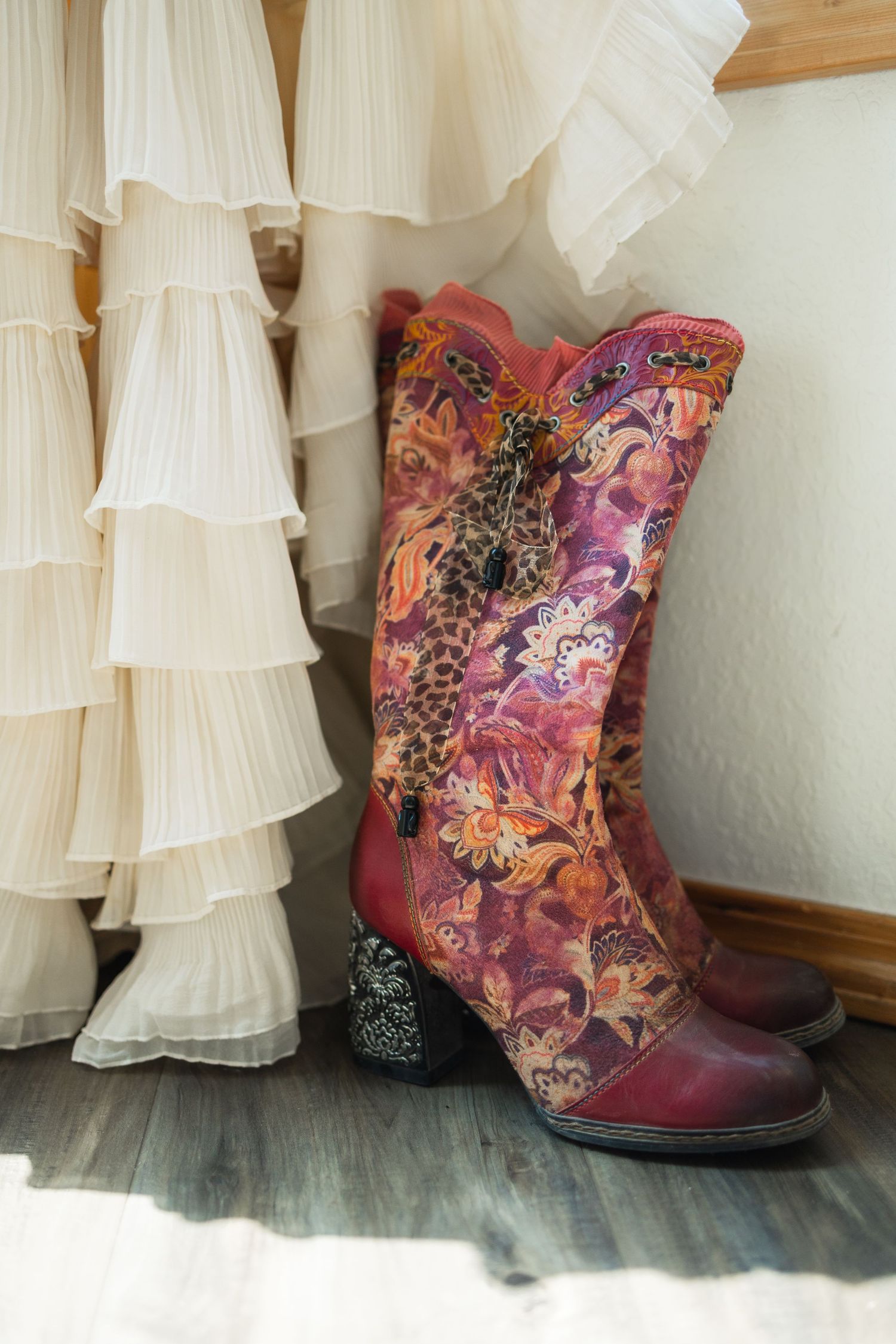 Colorful floral embroidered cowboy boots beside a ruffled white wedding dress on a wooden floor.