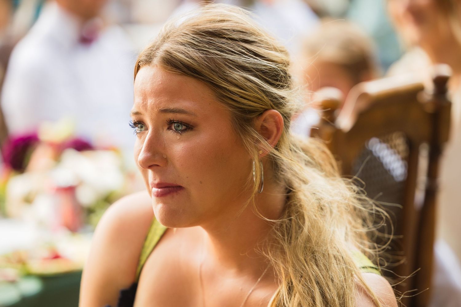 Emotional blonde woman with teary eyes in a yellow dress at an outdoor wedding reception.