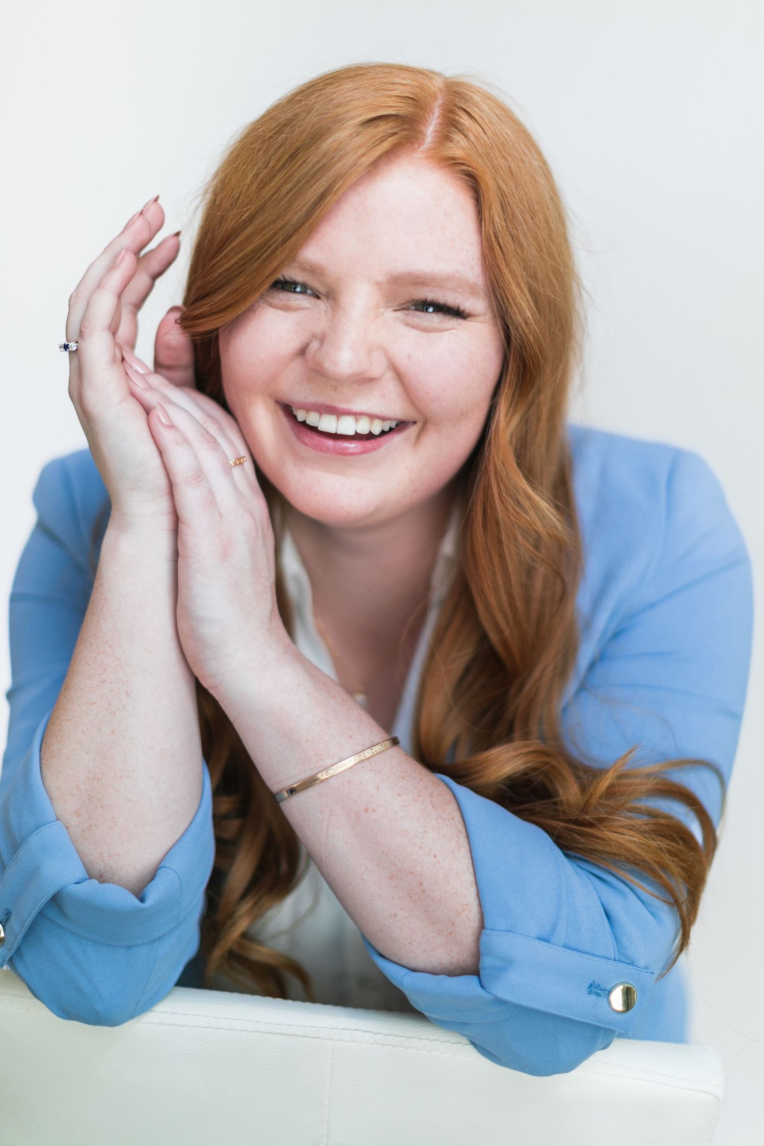 Smiling redhead woman with freckles in blue blazer, hands near face, wearing gold bracelet on white background.