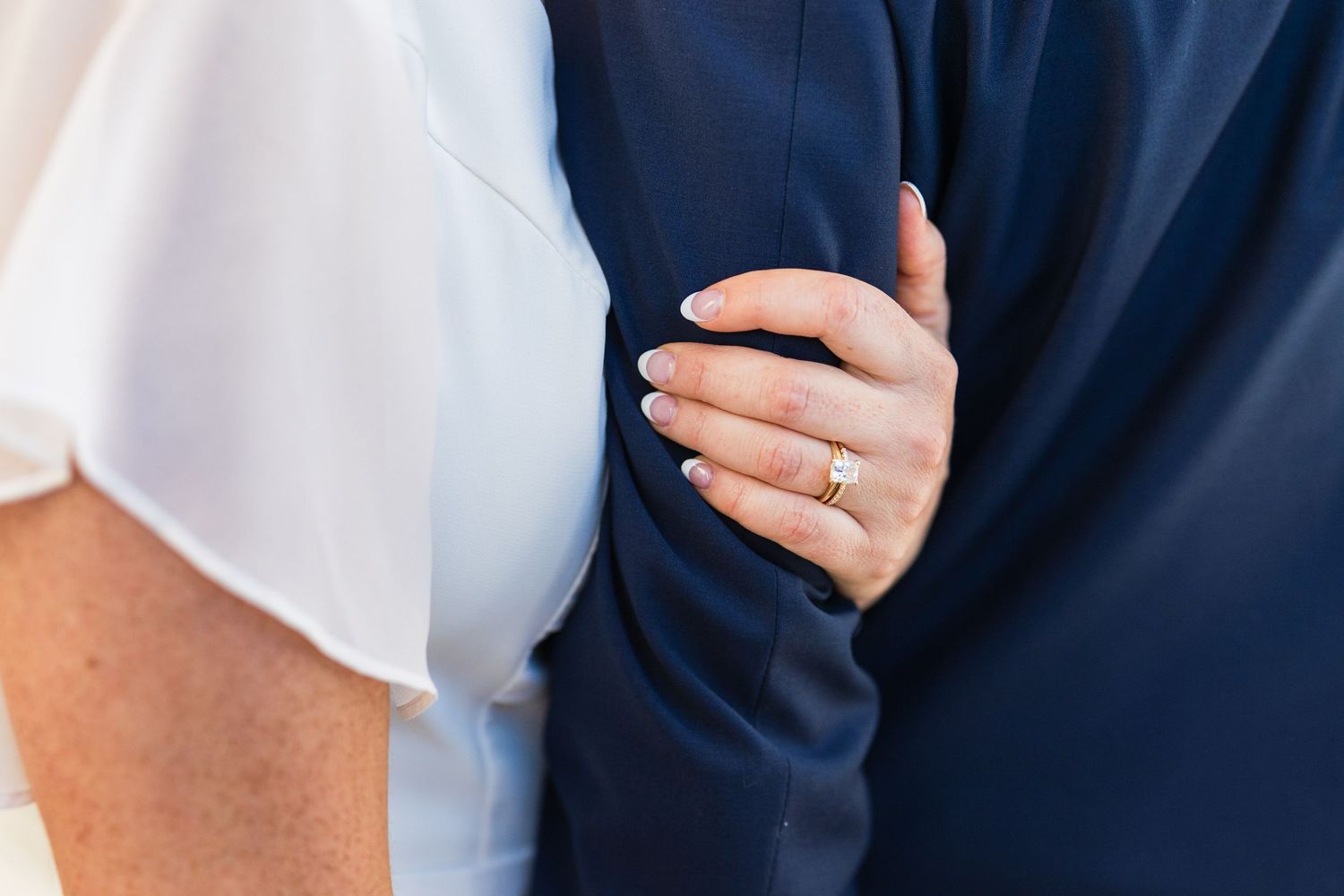 Close-up of bride's hand with diamond wedding ring resting on groom's navy blue suit jacket.