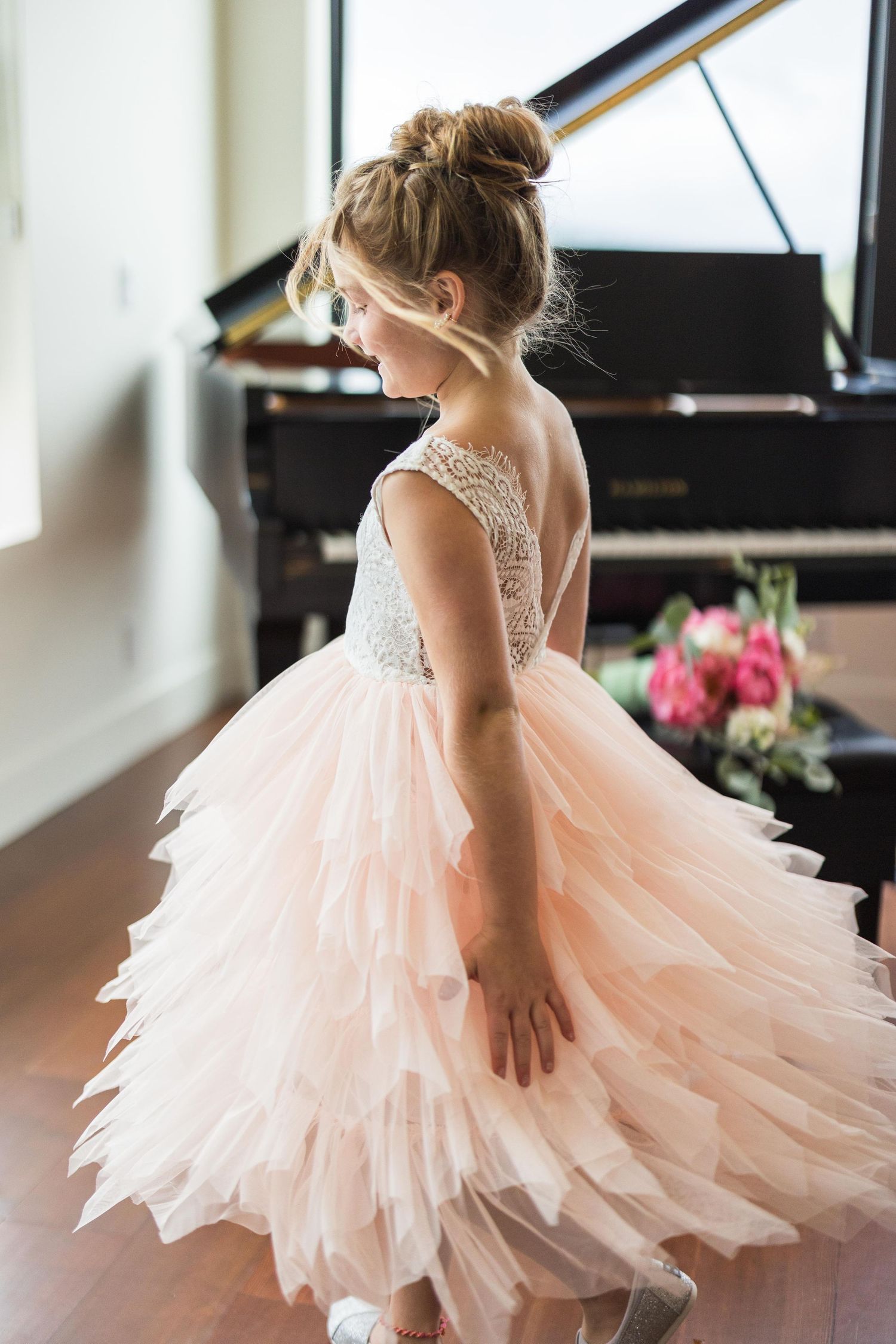 Young flower girl in layered blush pink tulle dress with lace bodice twirls near a grand piano.