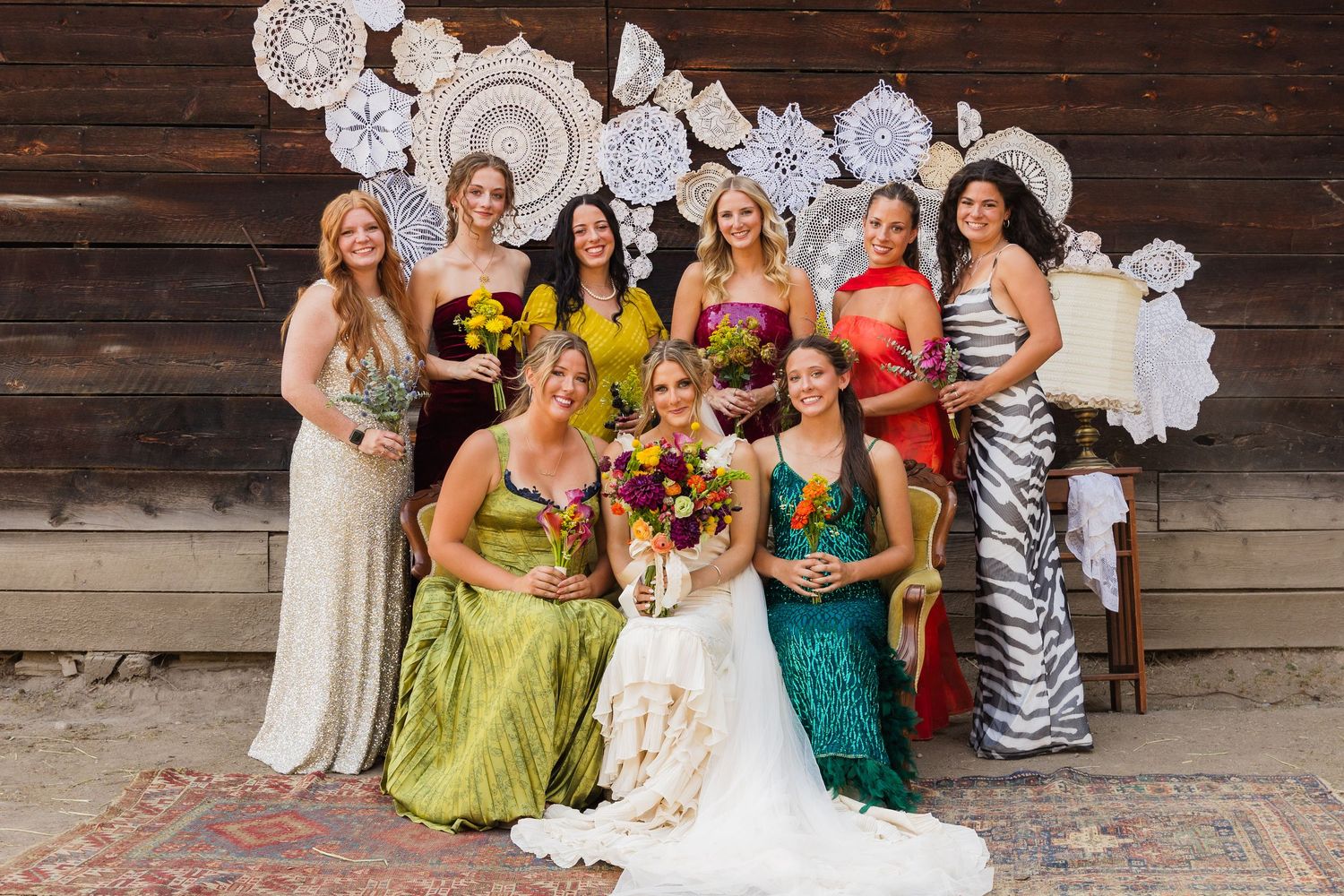 Bride in white gown poses with colorful bridesmaids in mismatched dresses before rustic wooden backdrop with doily decor.