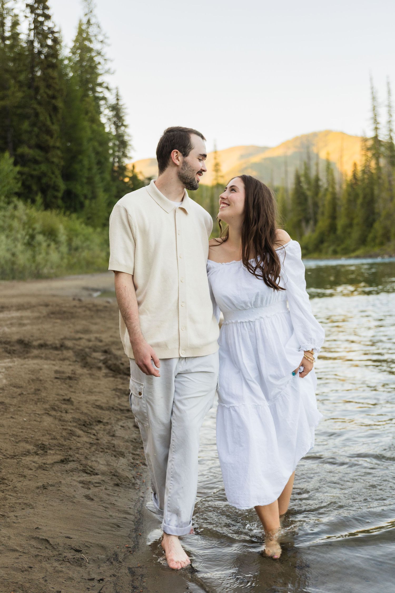 Couple walking barefoot along a mountain lake shore at sunset, woman in white dress, man in cream shirt and pants.