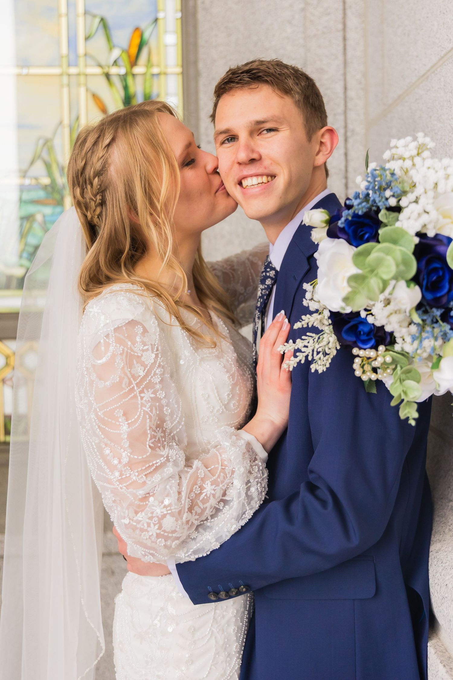 Bride kisses groom on the cheek as he smiles holding a blue and white bouquet indoors.