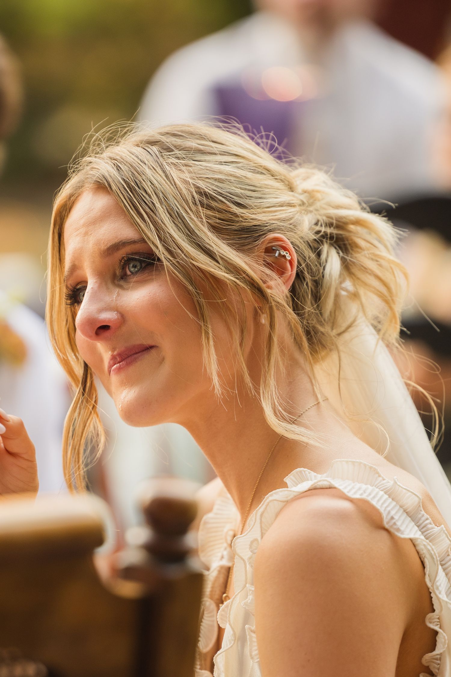 Emotional bride with updo hairstyle and ruffled white dress listens tearfully during outdoor wedding ceremony.