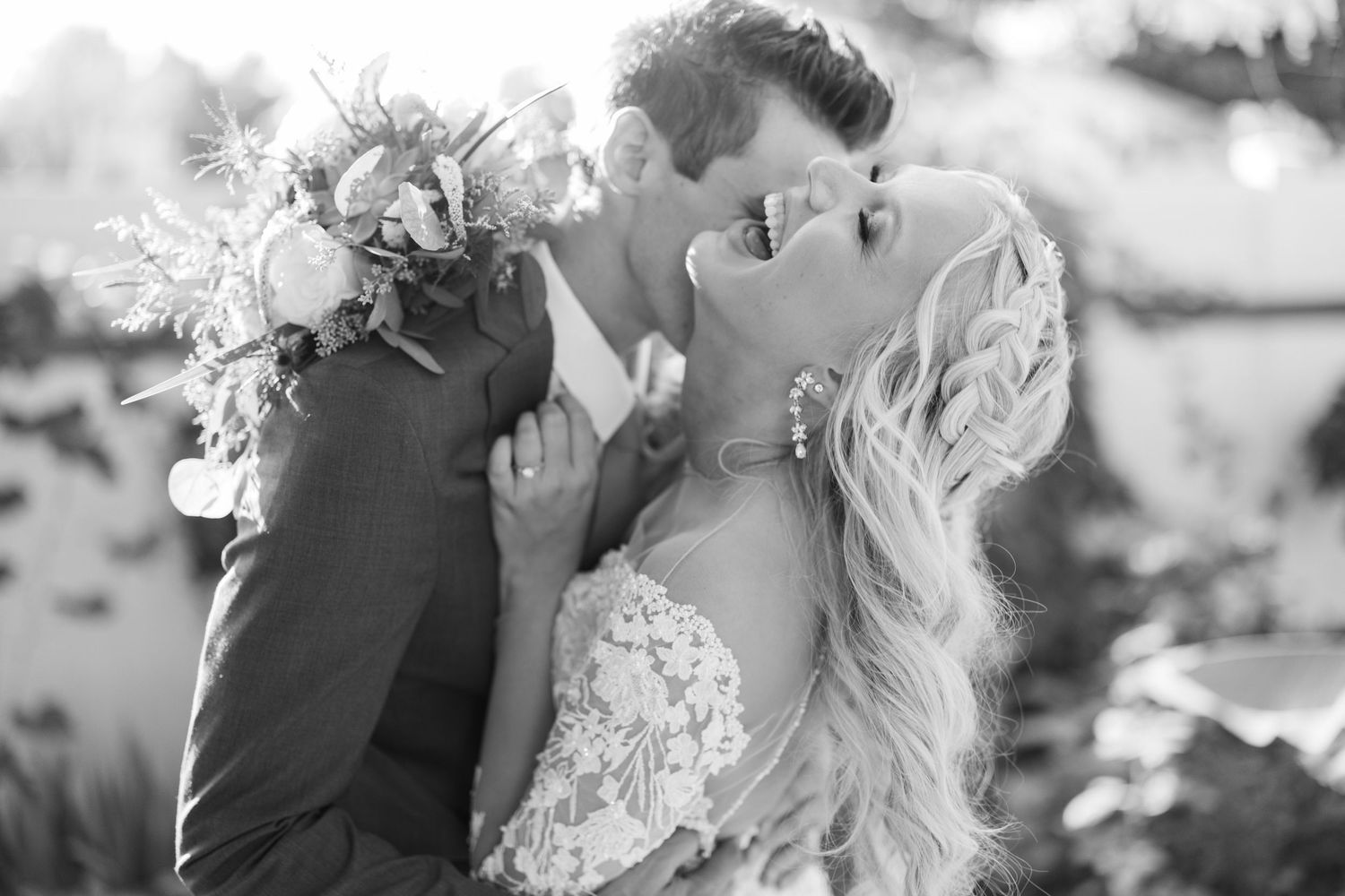 Bride and groom share romantic kiss with floral bouquet in black and white wedding portrait.