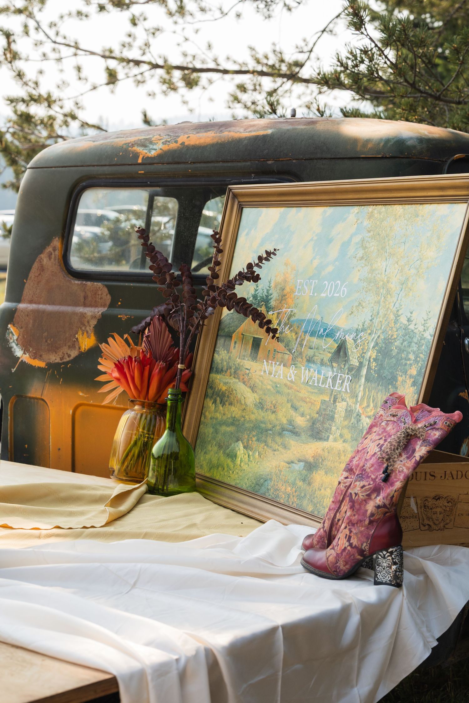 Rustic wedding welcome display with framed landscape painting, pink cowboy boots, and flowers on vintage truck bed.