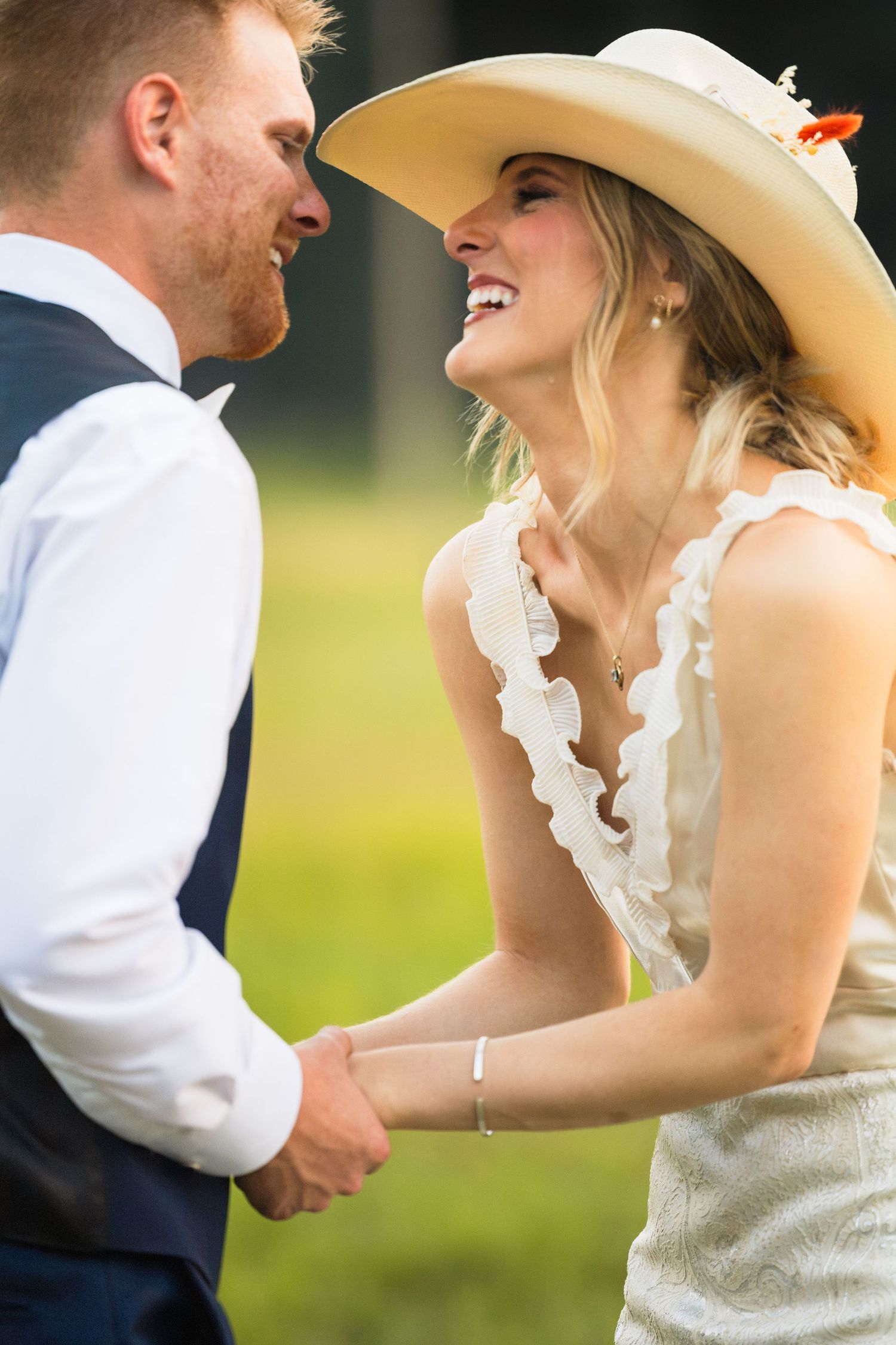 Bride in white ruffled dress and cowboy hat laughs joyfully while holding hands with groom in blue vest outdoors.