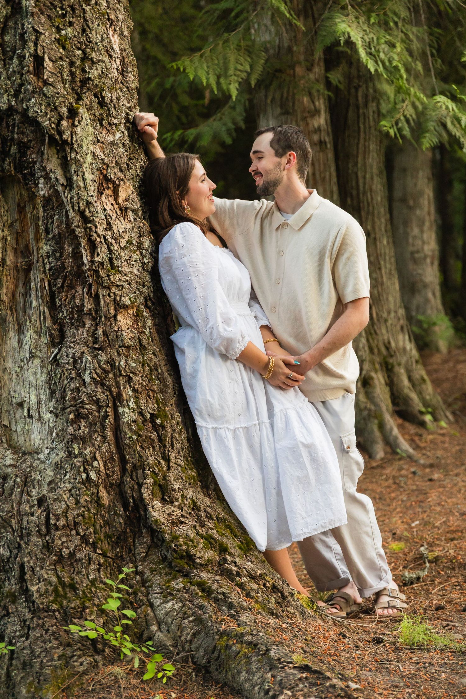 A couple in neutral tones share a romantic moment leaning against a large tree in a lush forest setting.
