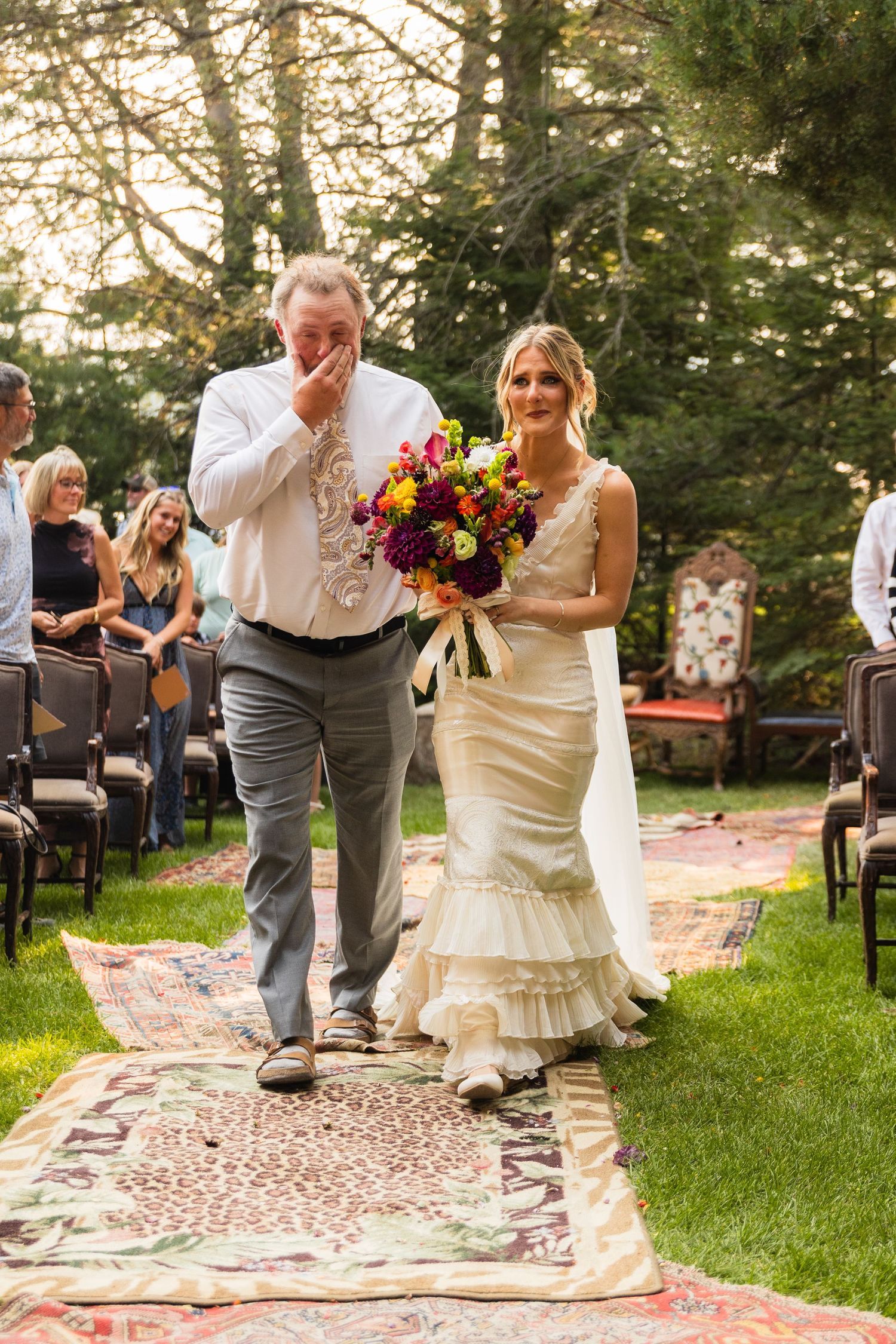 Bride in mermaid gown walks aisle with emotional father holding colorful bouquet at outdoor garden wedding ceremony.