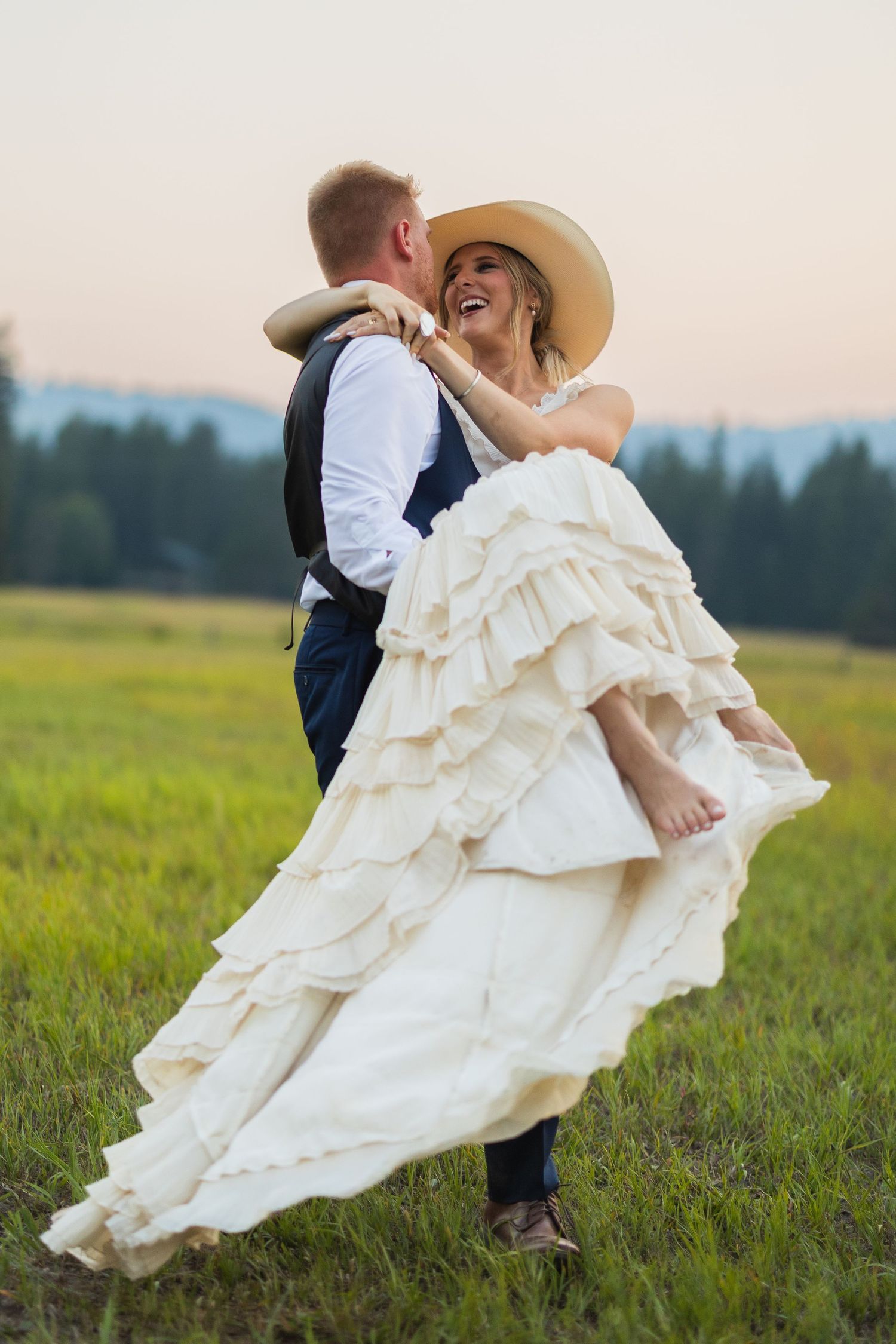 Groom lifts smiling bride in ruffled white dress and sun hat in a lush green field at sunset.